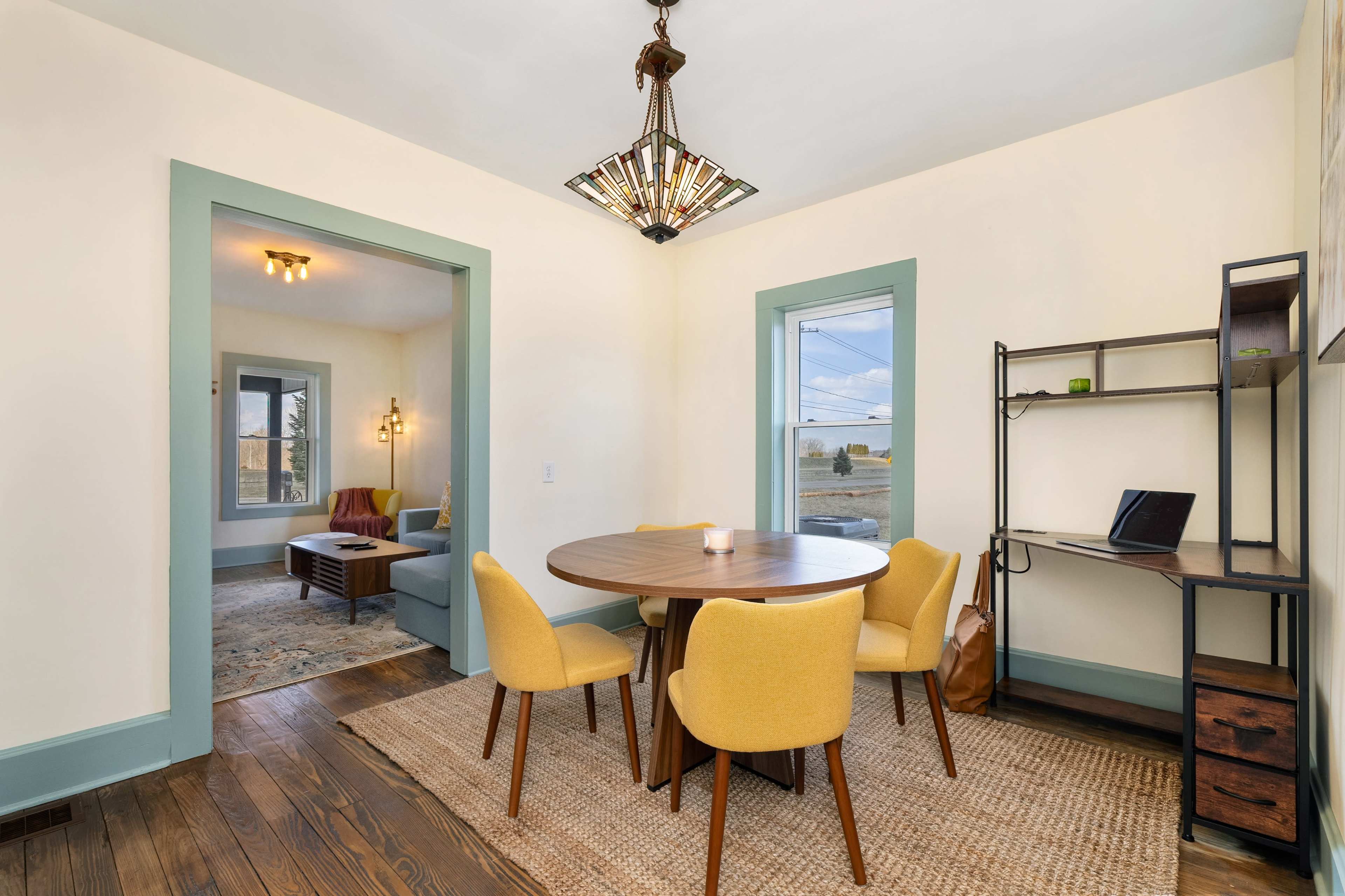 A dining area features a round table surrounded by yellow chairs, with a bookshelf and a window providing a view to the outside.