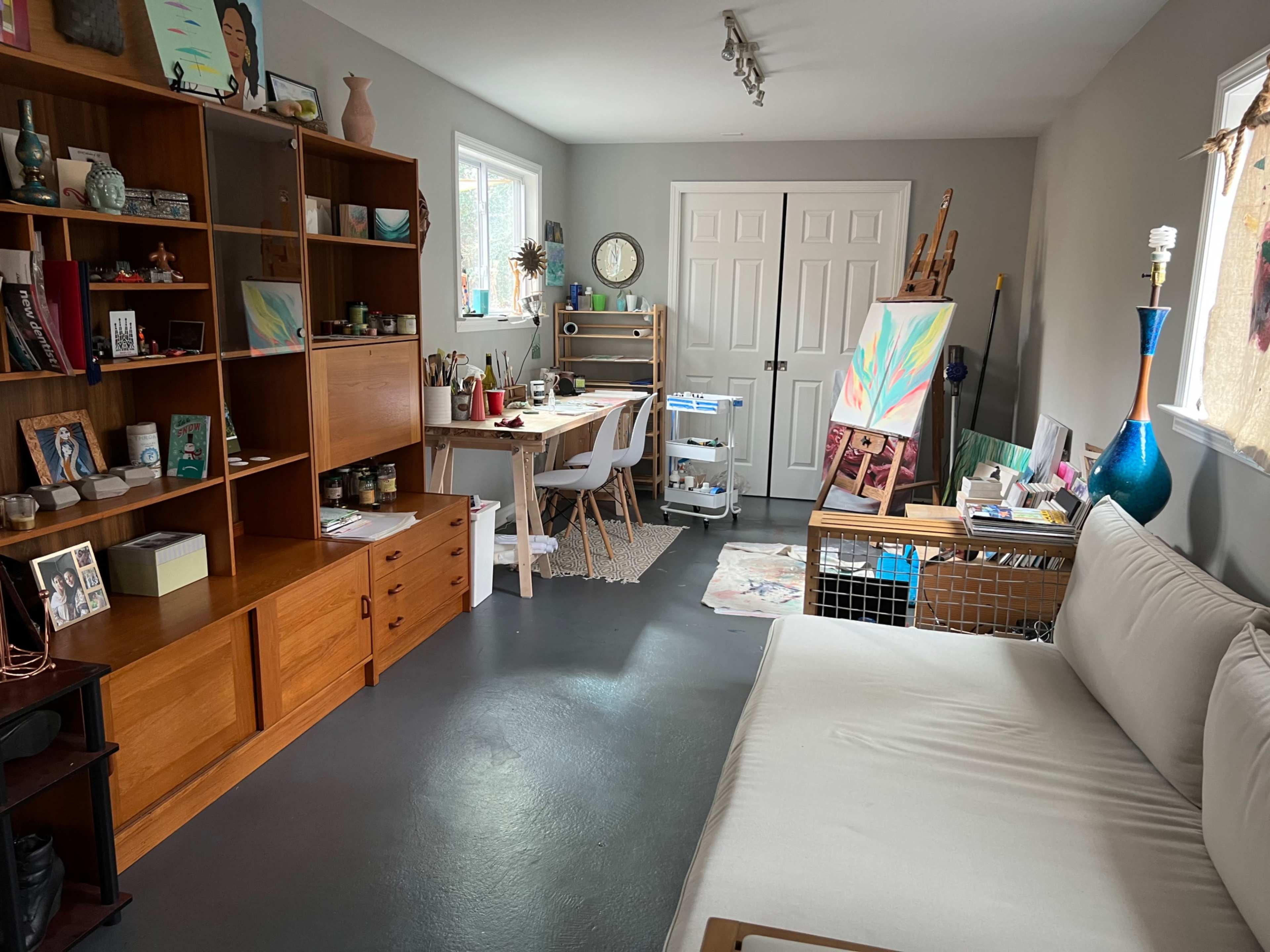 The image shows a cozy room with a sofa, wooden shelves filled with decorative items, a desk with art supplies, and an easel in one corner.