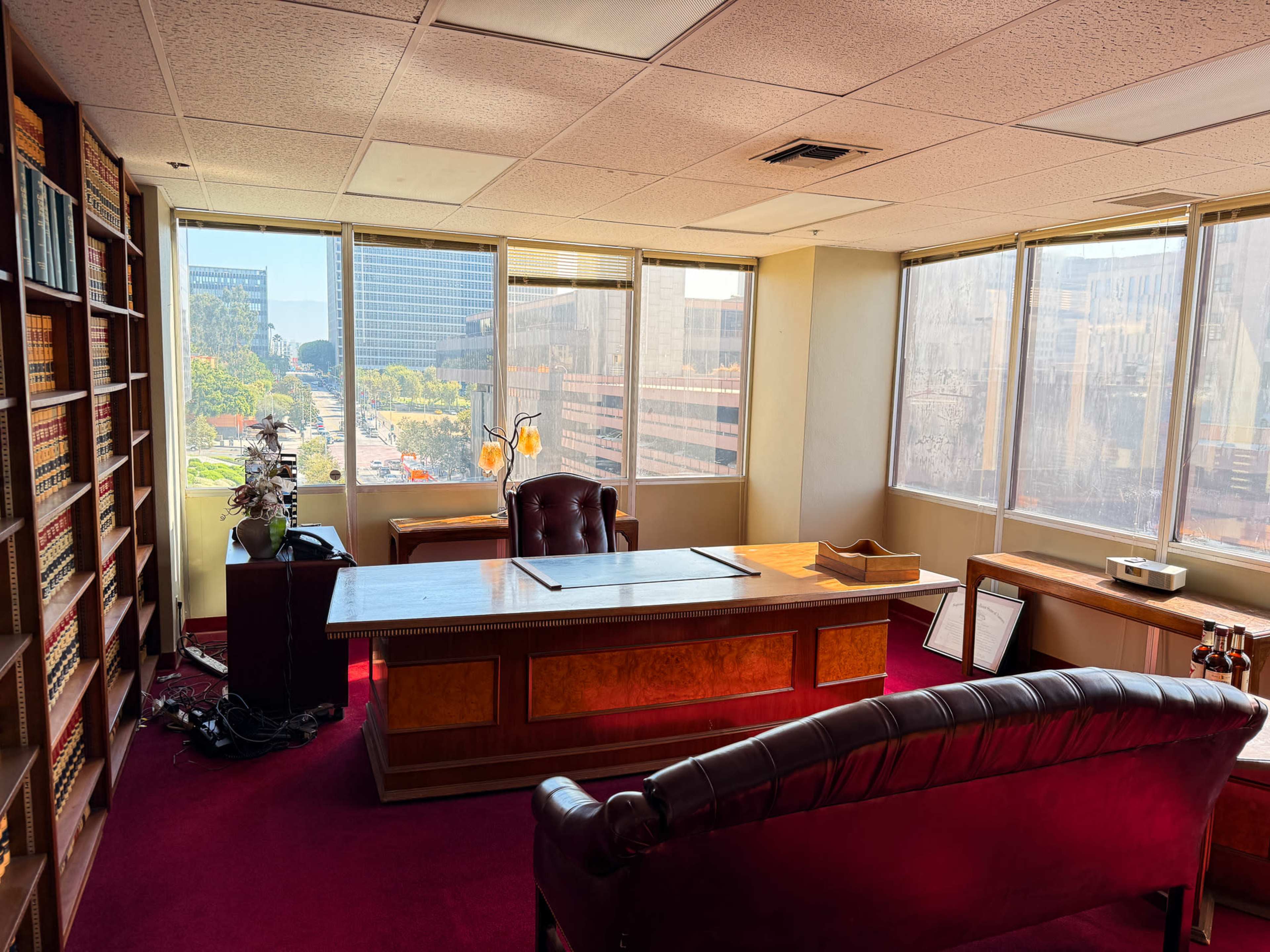The image shows a well-lit office with a large wooden desk, a leather chair, and bookshelves filled with books, overlooking a cityscape through large windows.