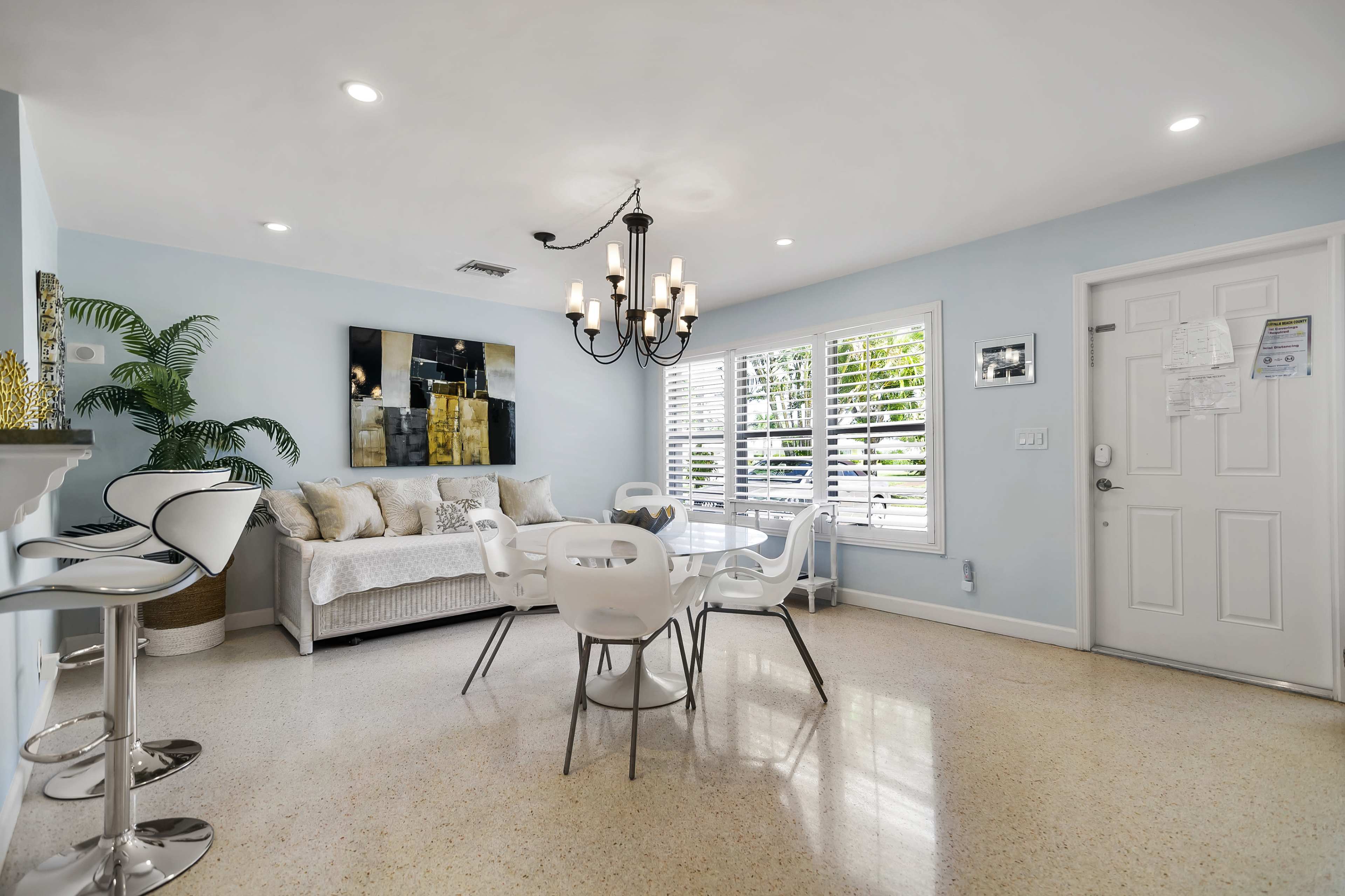 A dining area features a round table with white chairs, a chandelier, a gray sofa, and a large window with shutters.