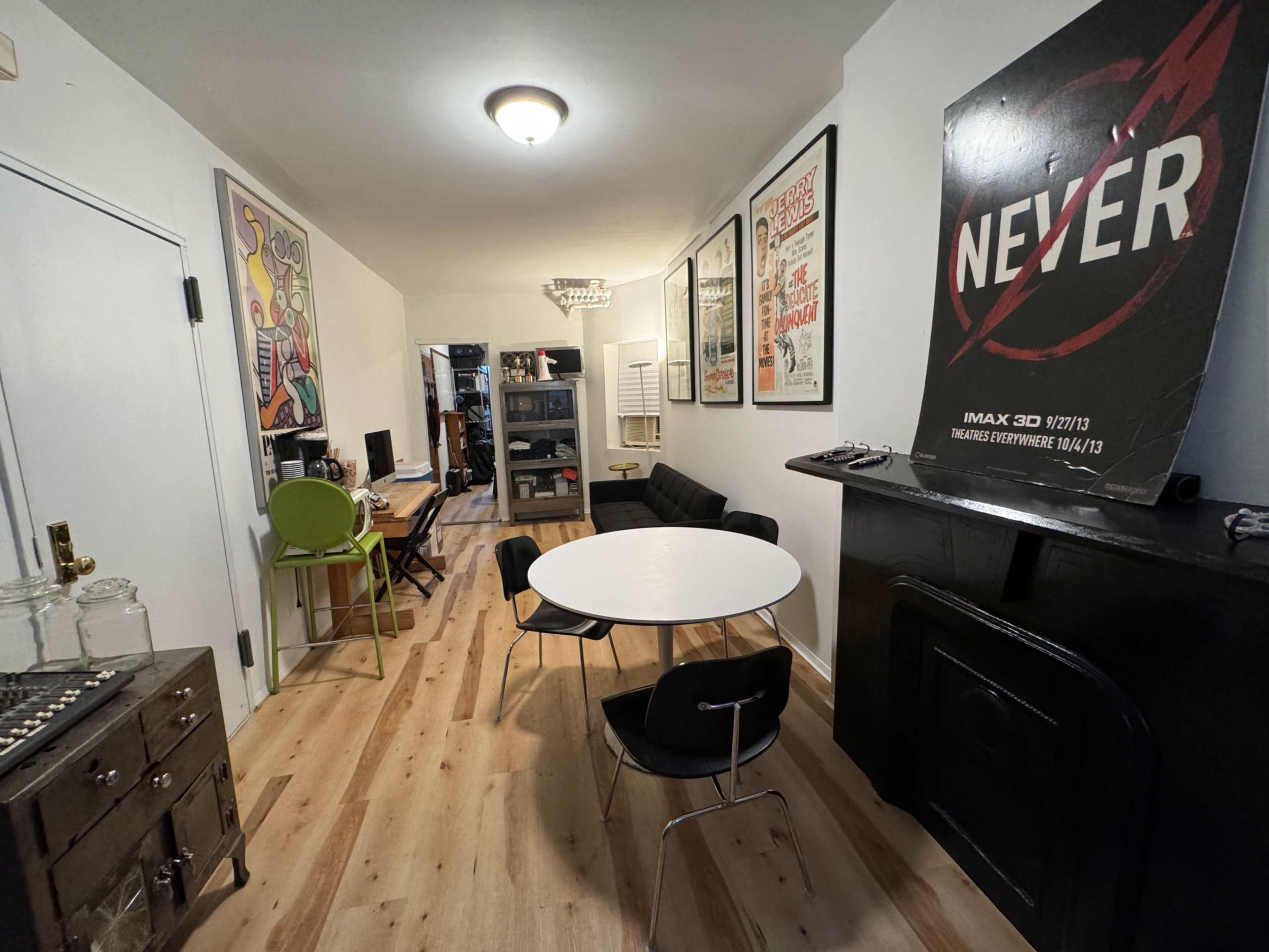 The image shows a narrow room with light-colored wooden flooring, featuring a round white dining table surrounded by chairs, modern artwork on the walls, and a black fireplace.