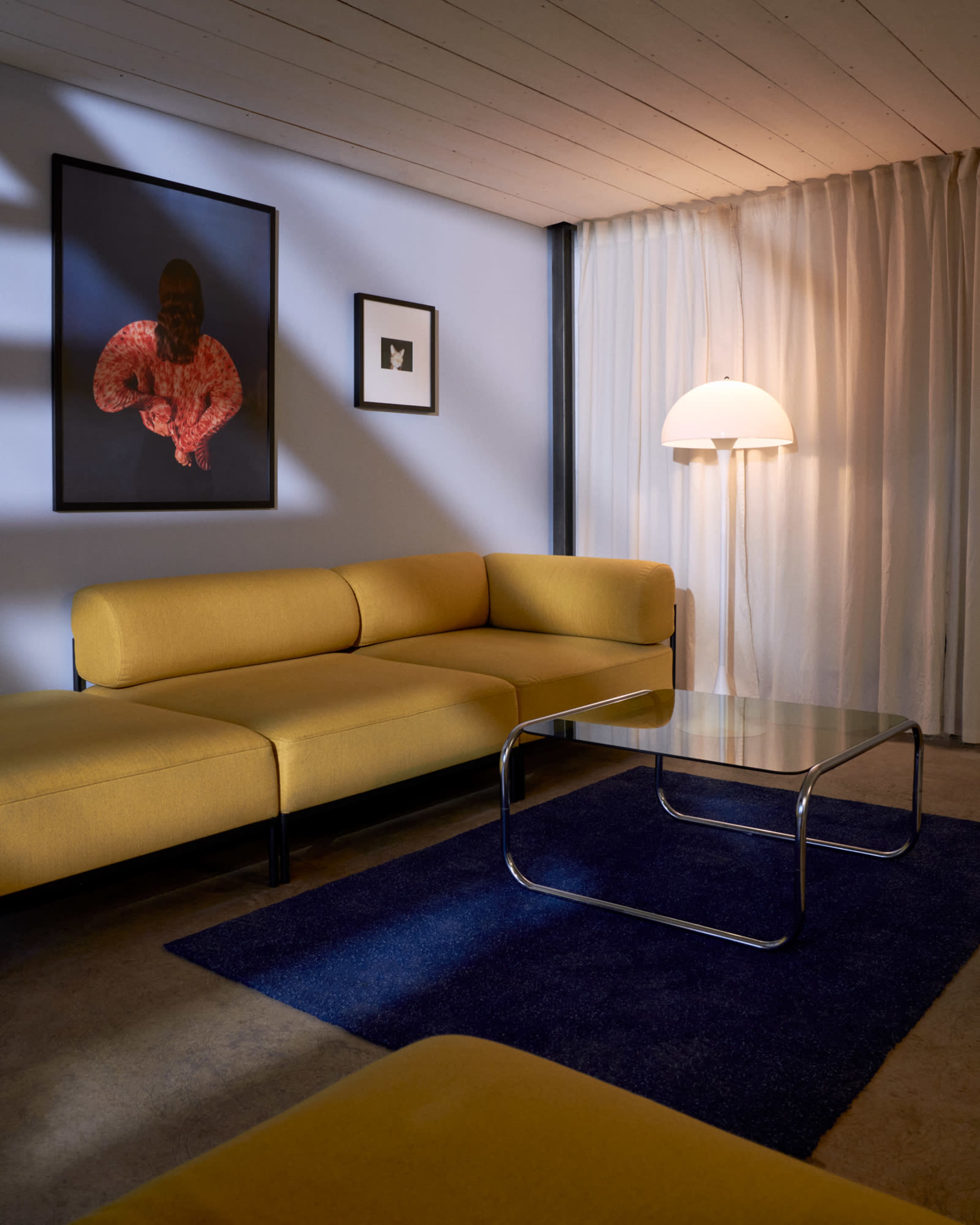 A modern living room features a yellow sectional sofa, a glass coffee table, a blue area rug, and an art piece on the wall, illuminated by a floor lamp.