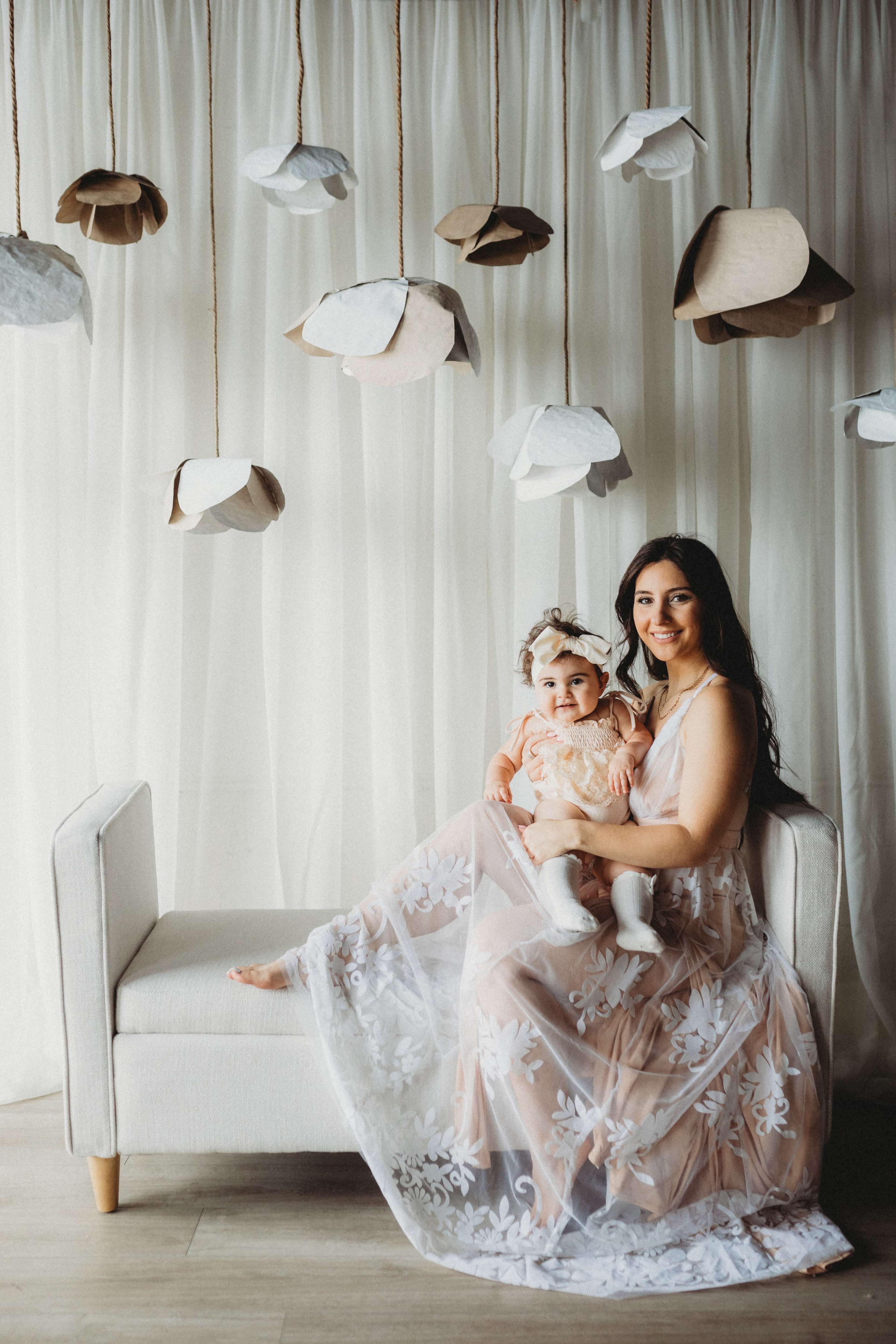 A woman in a light dress sits on a sofa holding a baby, surrounded by hanging paper flowers against a white backdrop.