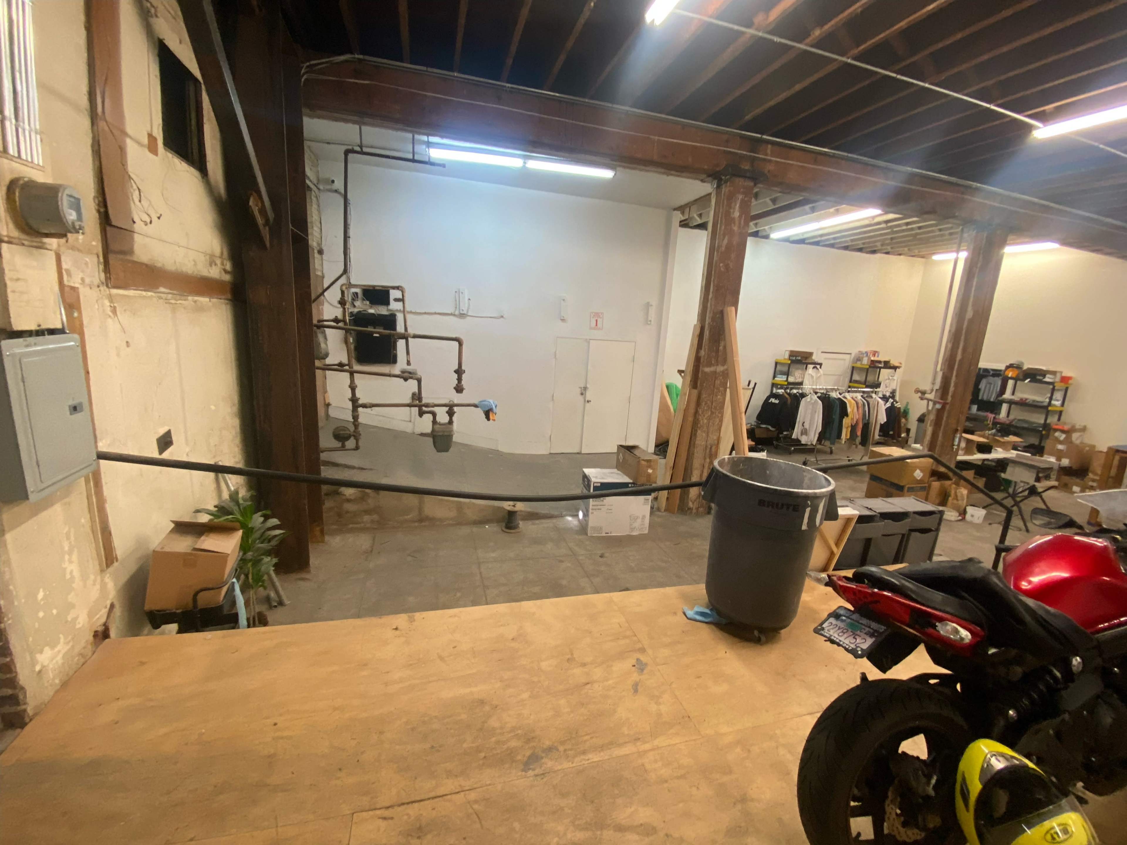 A spacious, unfinished industrial storage area with wooden beams, concrete floor, and various items stacked around, including a motorcycle and boxes.