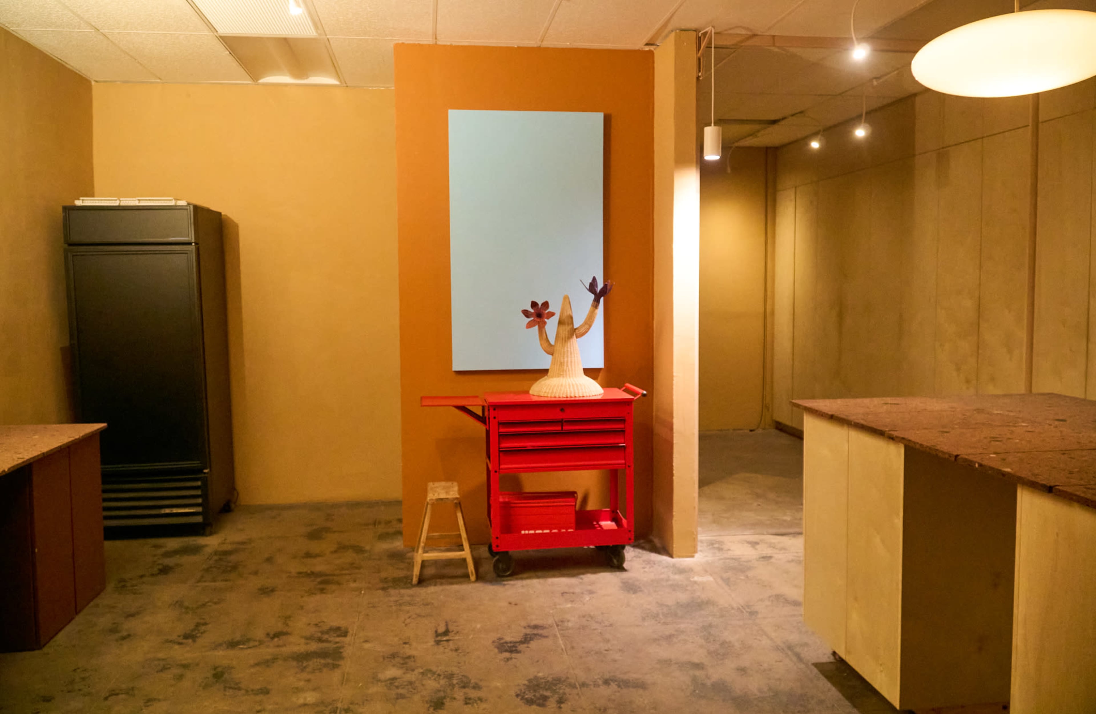 An empty, well-lit room features two work tables, a red cart with a decorative object, a large mirror on the wall, and a black refrigerator in the corner.