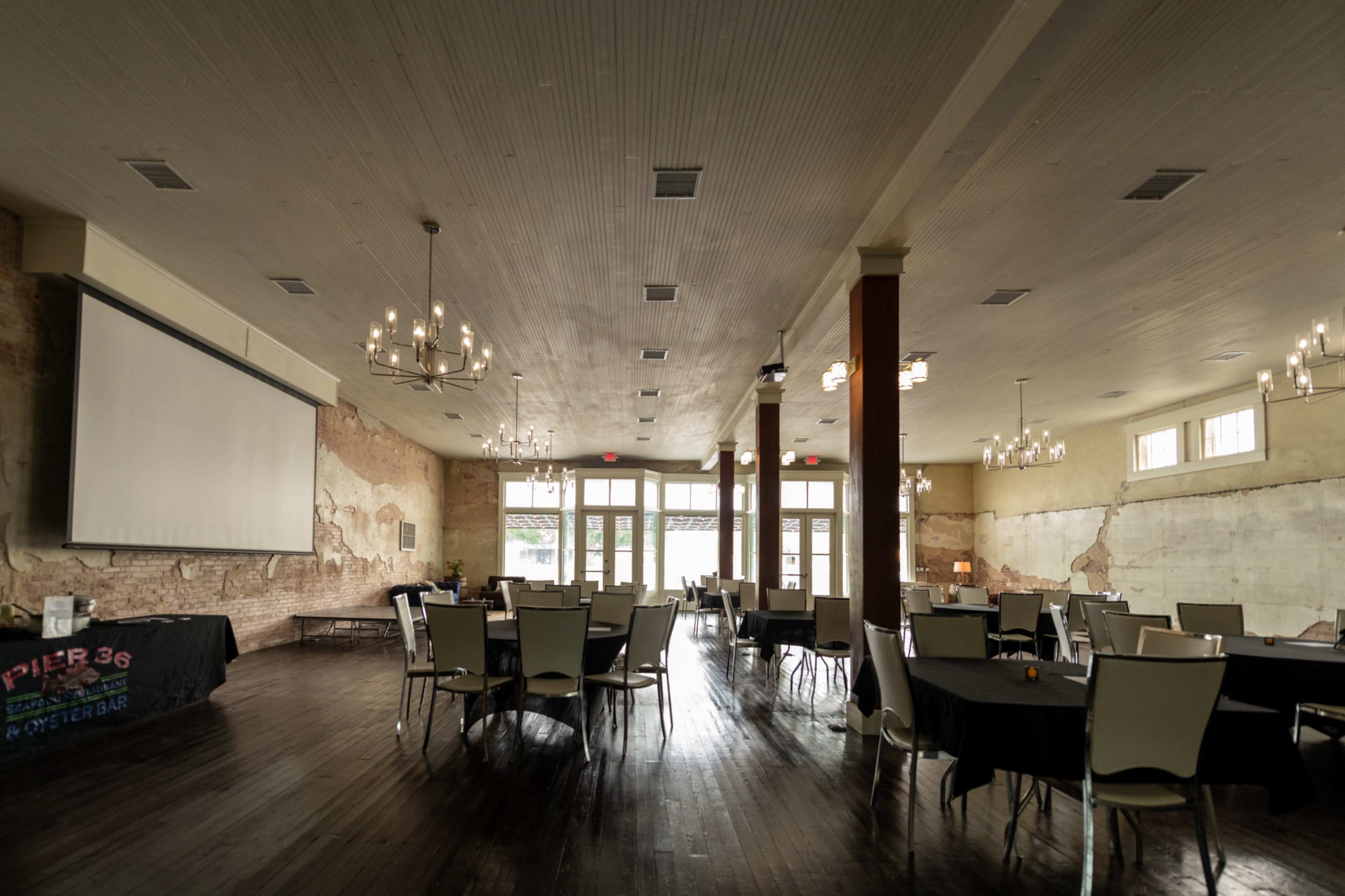 The image shows a spacious indoor event venue with tables set for dining and a large projector screen mounted on a wall.