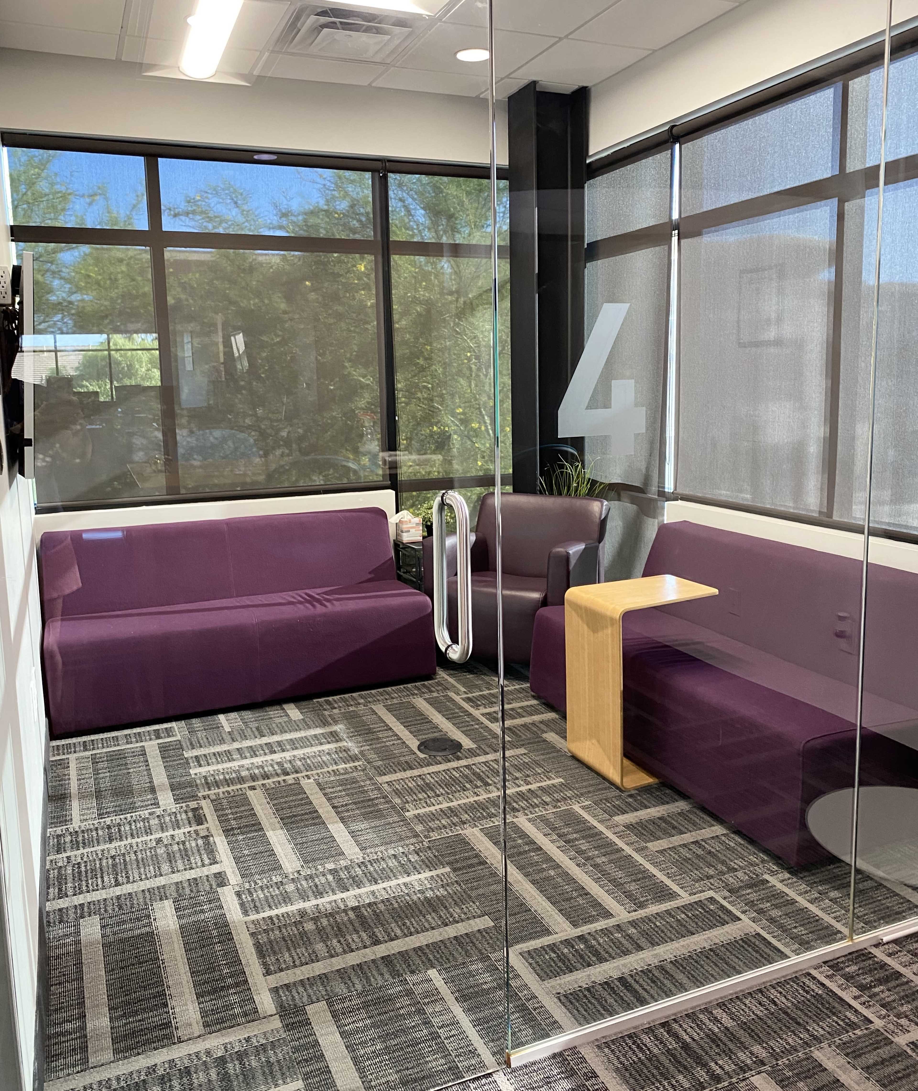 A small meeting room with two purple sofas, a wooden table, and a chair, surrounded by large windows letting in natural light.