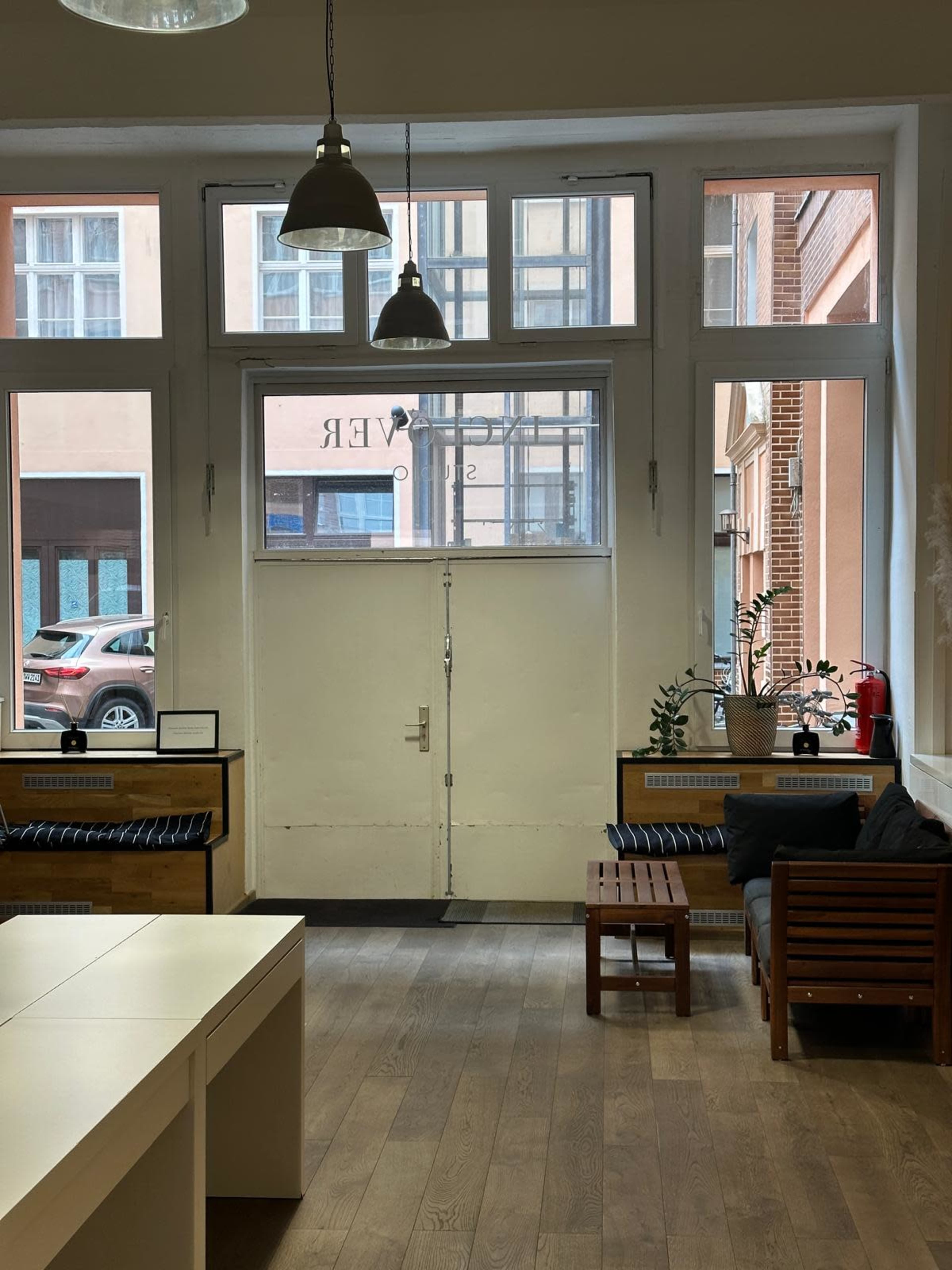 The image shows a bright room with large windows, an entrance door, wooden flooring, and minimalist furniture including a sofa and small tables.