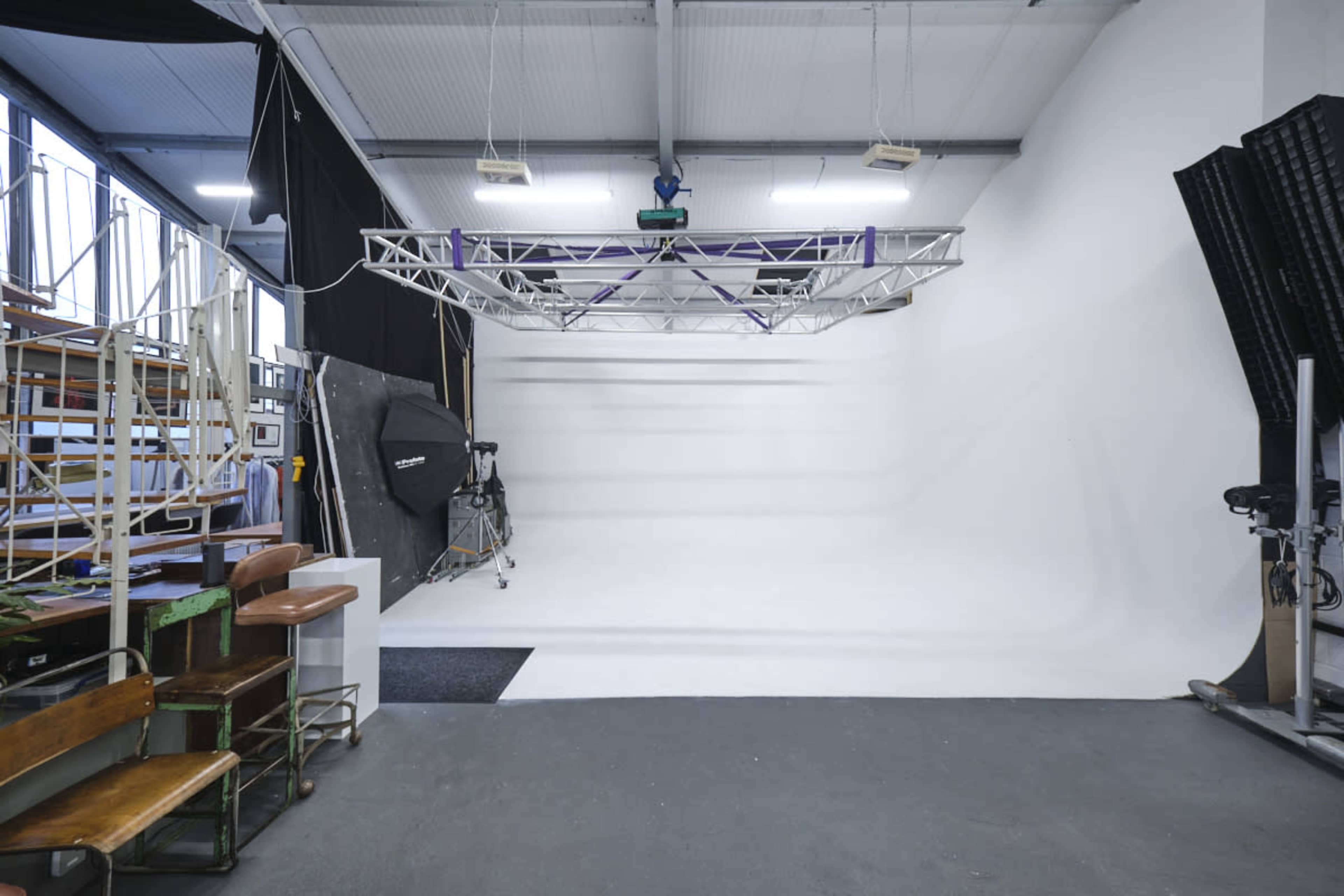 A spacious photography studio with a white background, a lighting rig overhead, and various equipment along the sides.