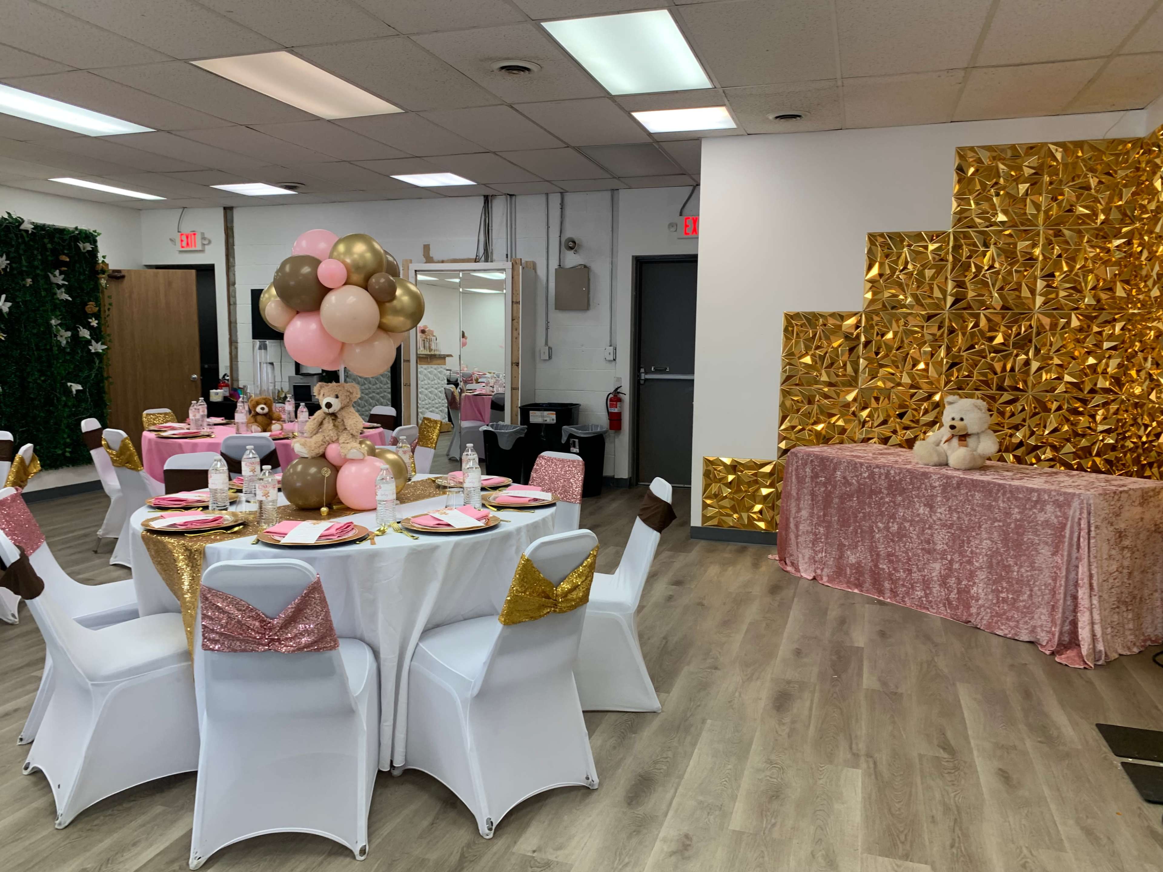 A decorated party room with tables set in white and gold, featuring balloon centerpieces and a gold backdrop.