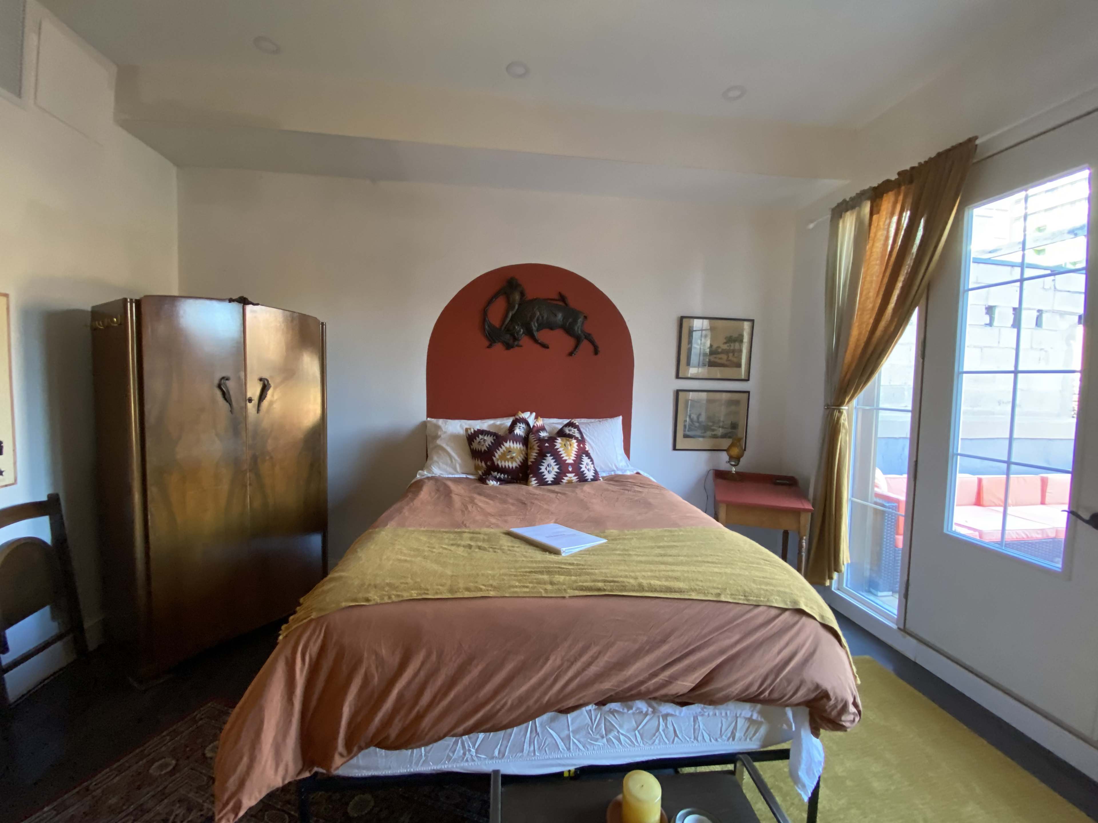 A cozy bedroom featuring a bed with an orange-brown cover, a wooden wardrobe, and a small table by a window.