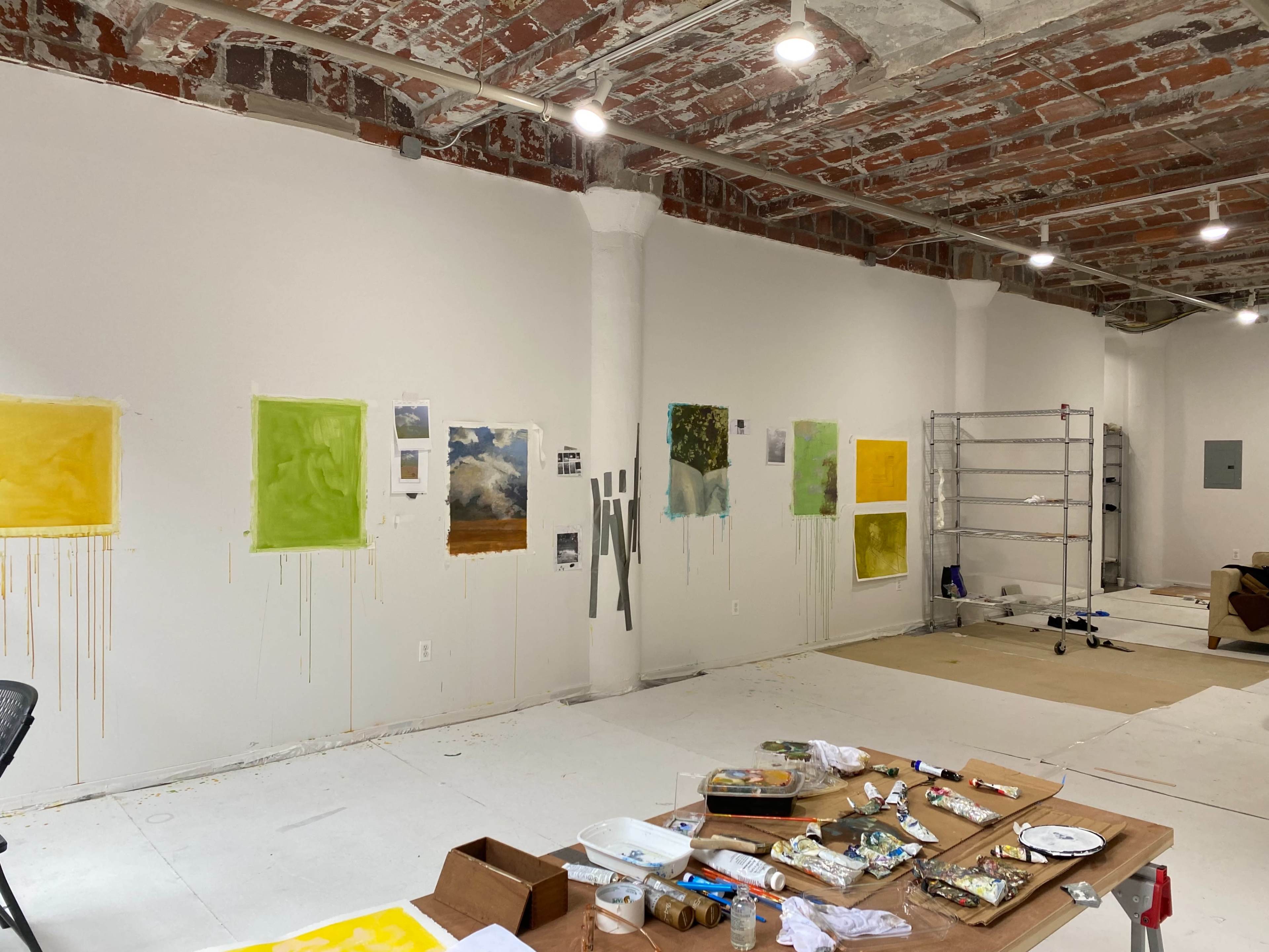 A spacious, unfinished art studio with paint-splattered walls and a table covered in art supplies, alongside several artworks displayed in various shades of yellow and green.