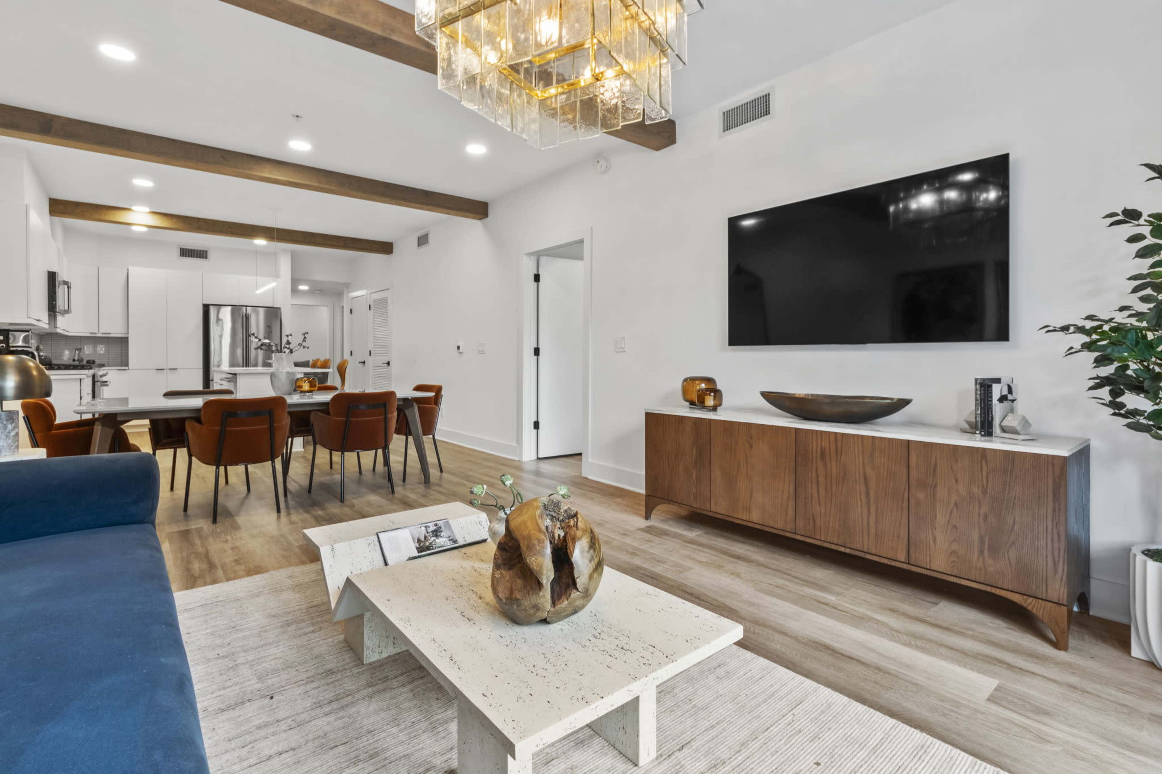 A modern living room features a blue sofa, a wooden coffee table, a large television mounted on the wall, and a dining area with a table and chairs in the background.