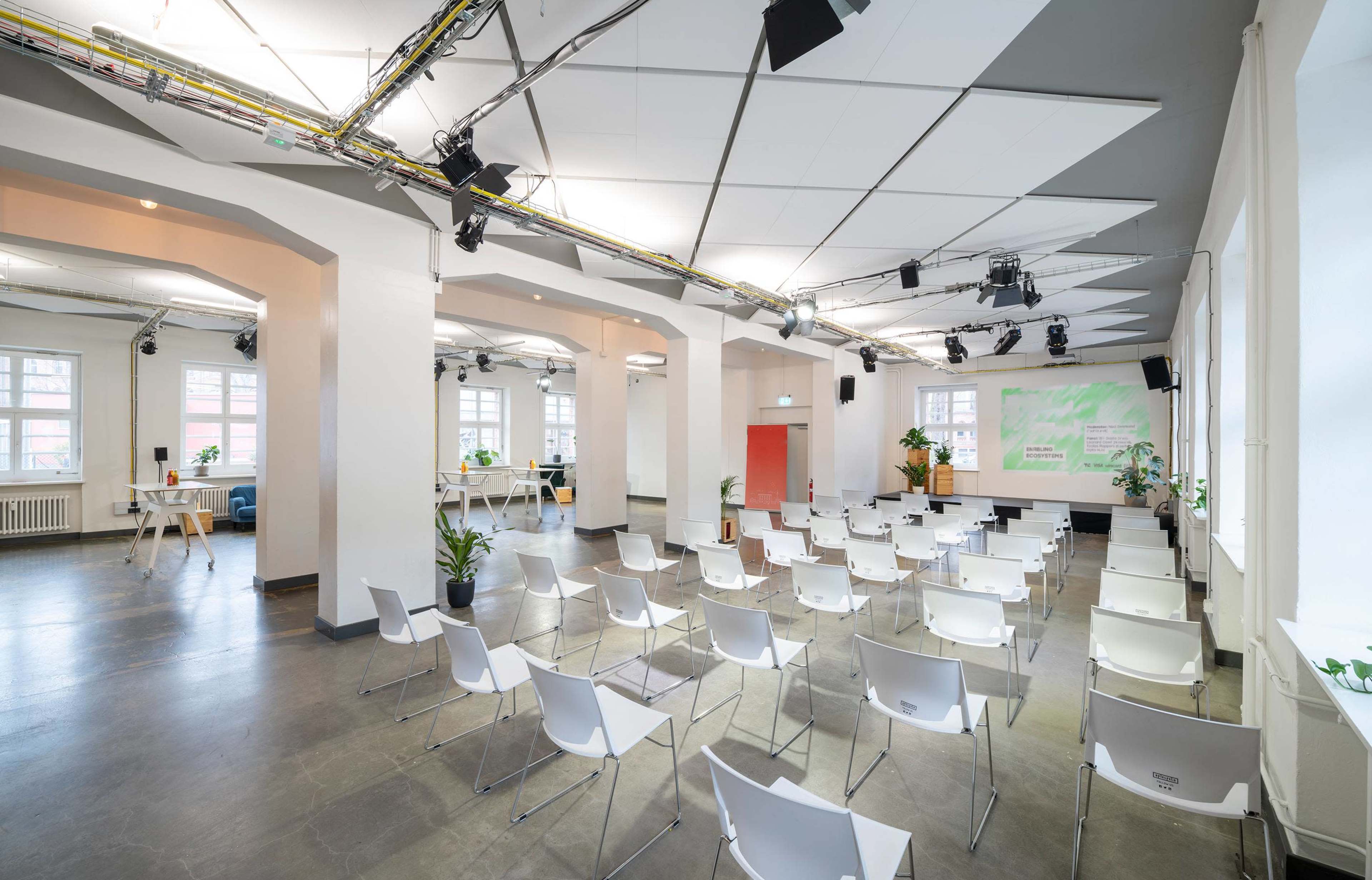 A spacious seminar room is set up with empty white chairs arranged in rows facing a projected screen, surrounded by plants and bright lighting.