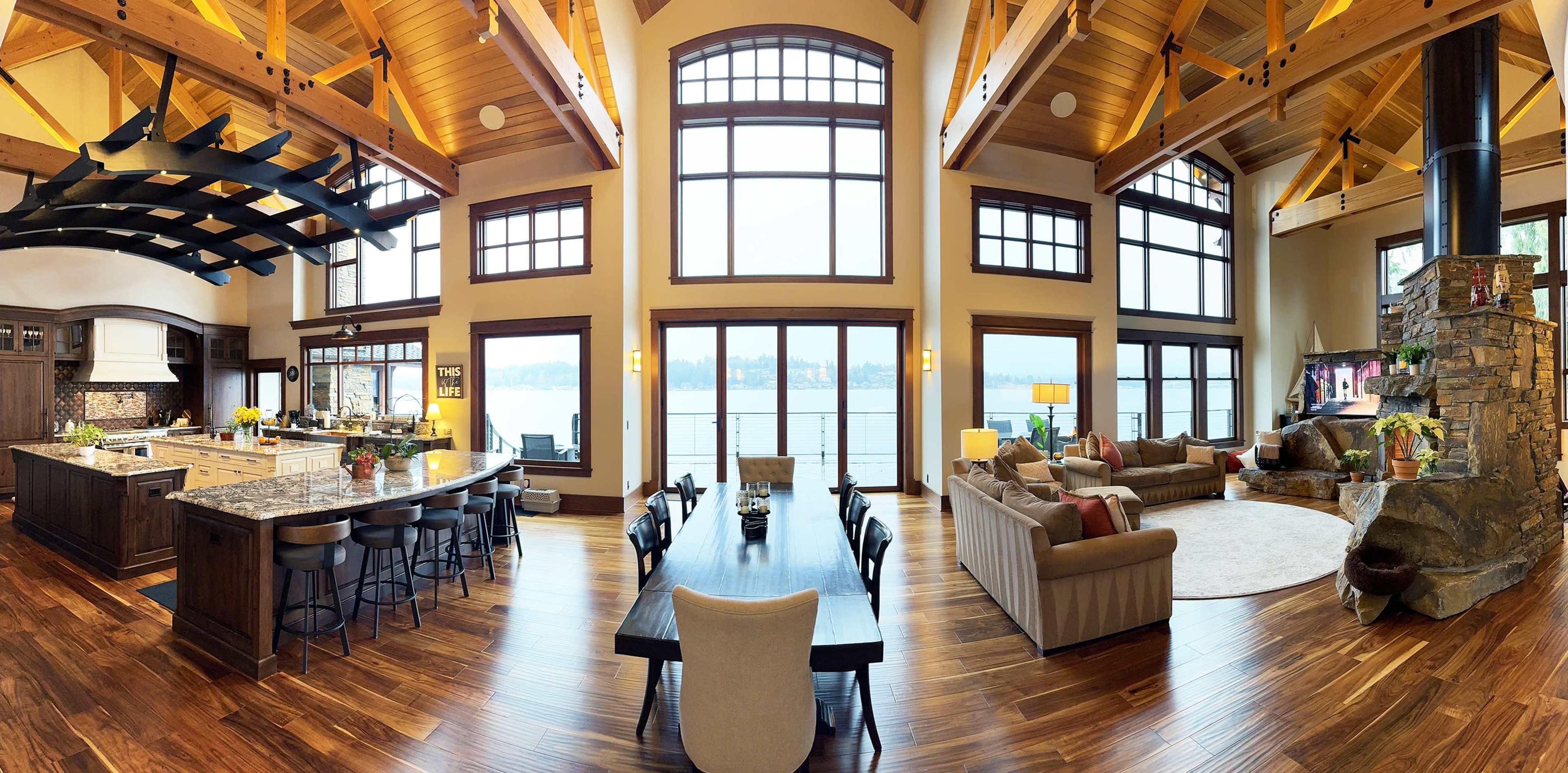 A panoramic view of a spacious open-concept living area featuring a kitchen, dining area, and a cozy seating arrangement next to large windows overlooking a body of water.