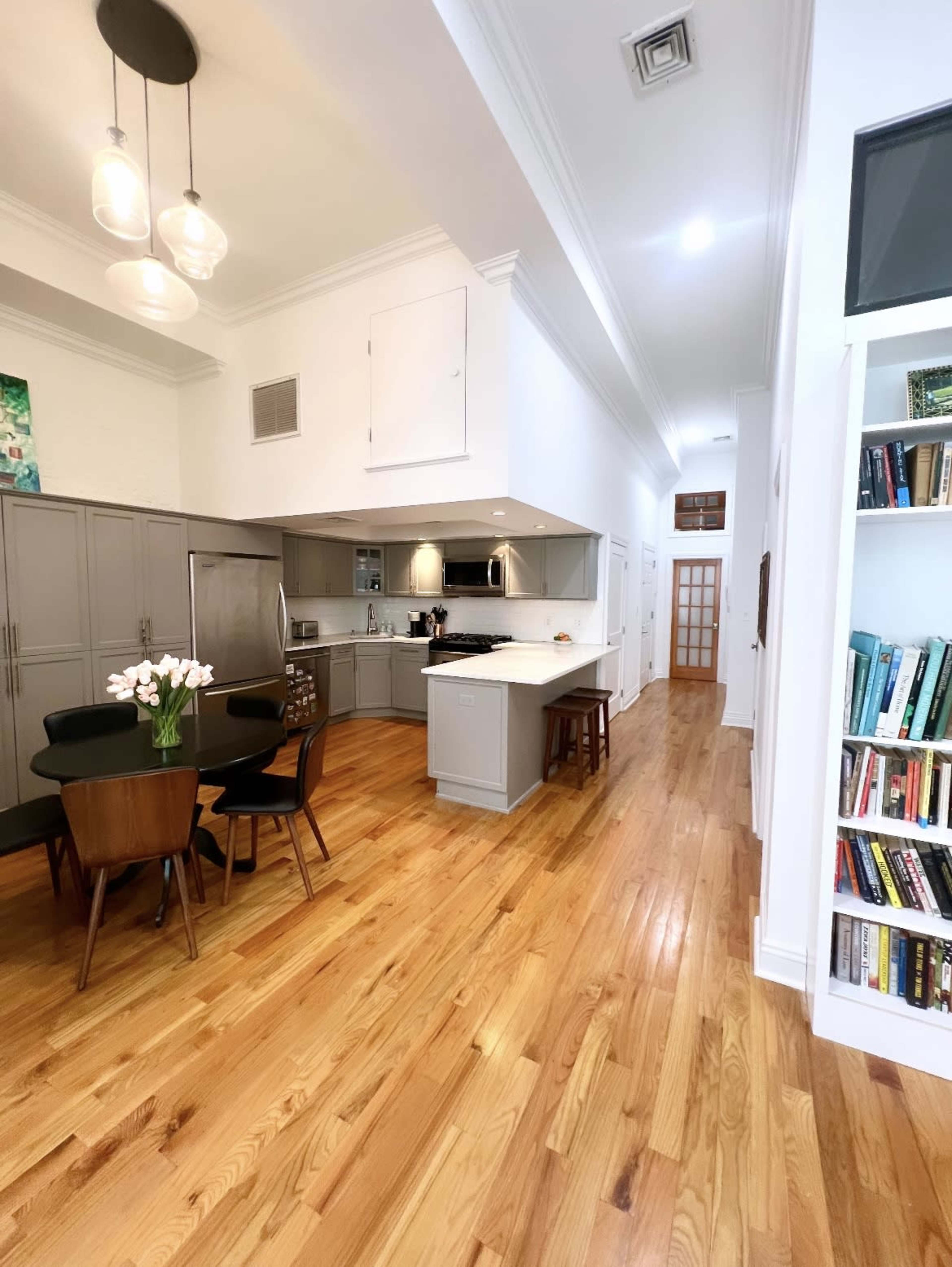 A modern kitchen and dining area with wooden flooring, a large island, and a bookshelf filled with books along one wall.