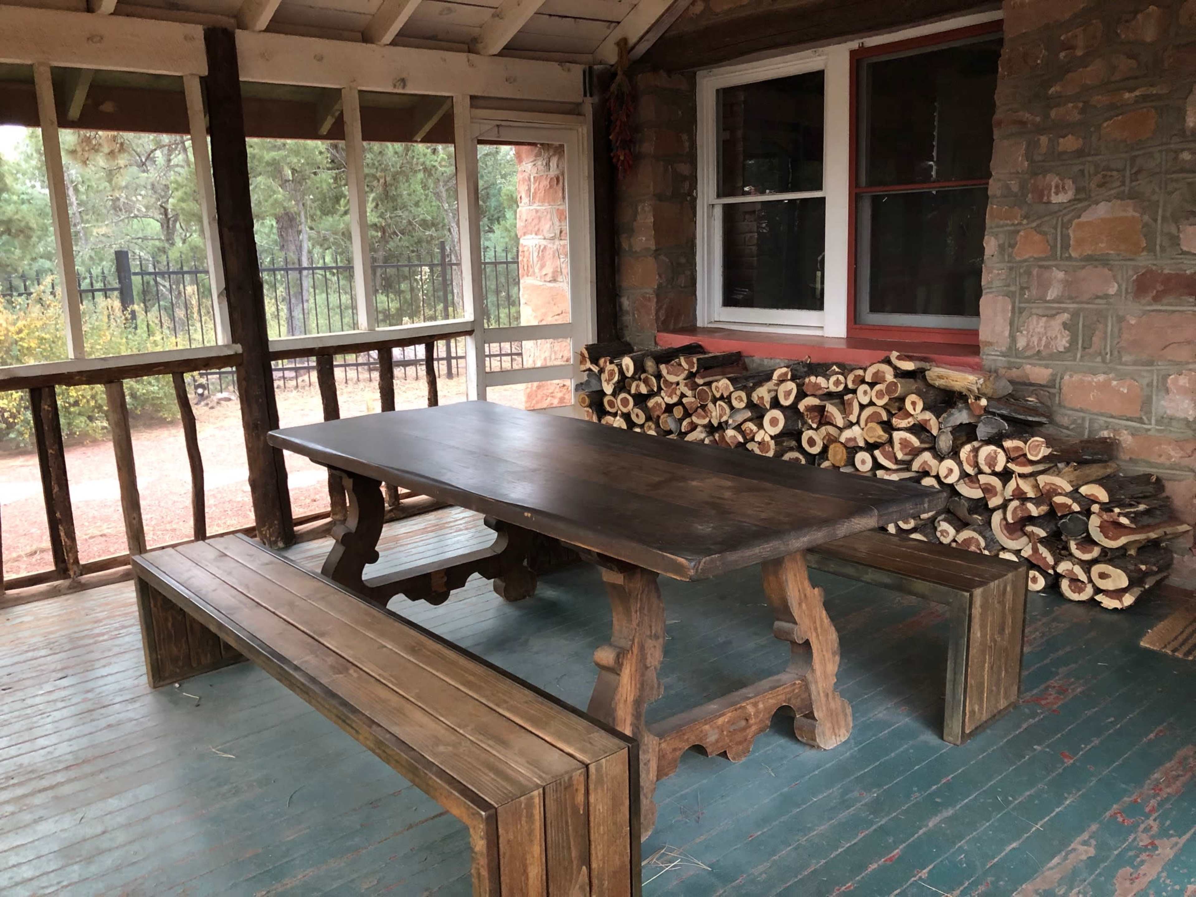 A wooden table and benches are positioned on a porch next to a stack of firewood against a stone wall.