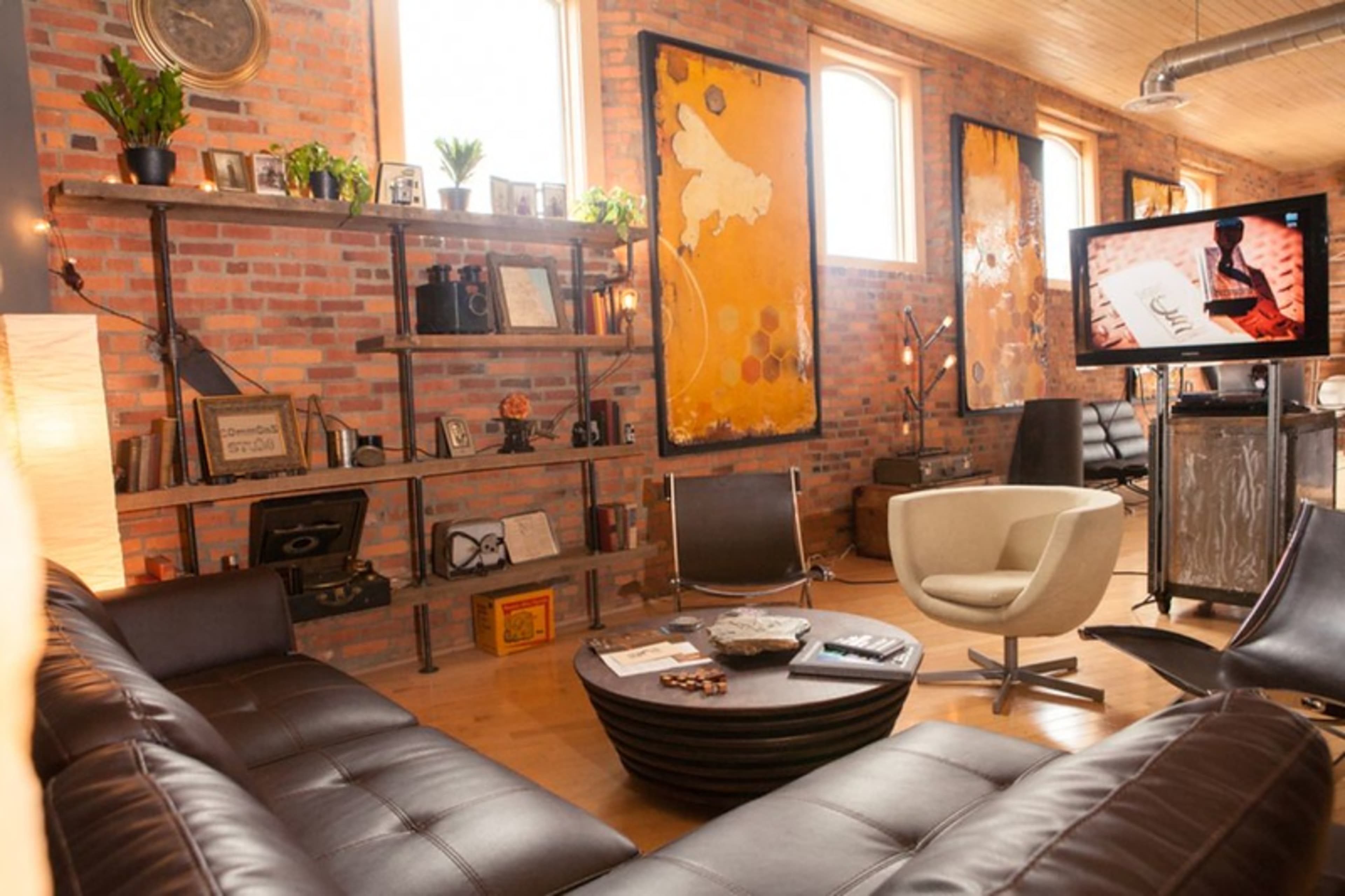 A contemporary living room features a sectional leather sofa, a round coffee table, a television, and brick walls adorned with artwork and shelves displaying plants and decorative items.