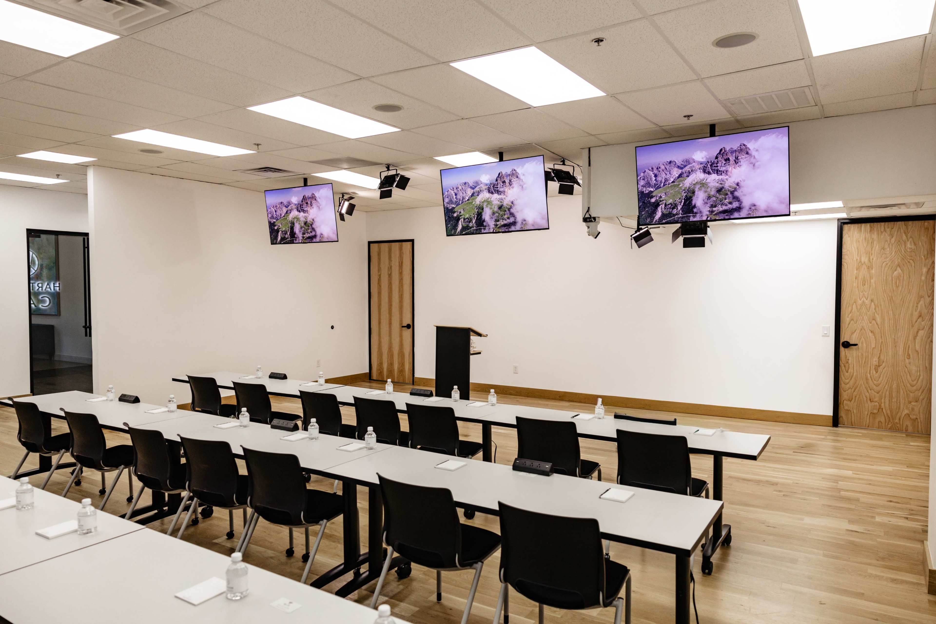 The image shows a conference room with rows of tables and chairs facing three mounted screens displaying mountain landscapes.