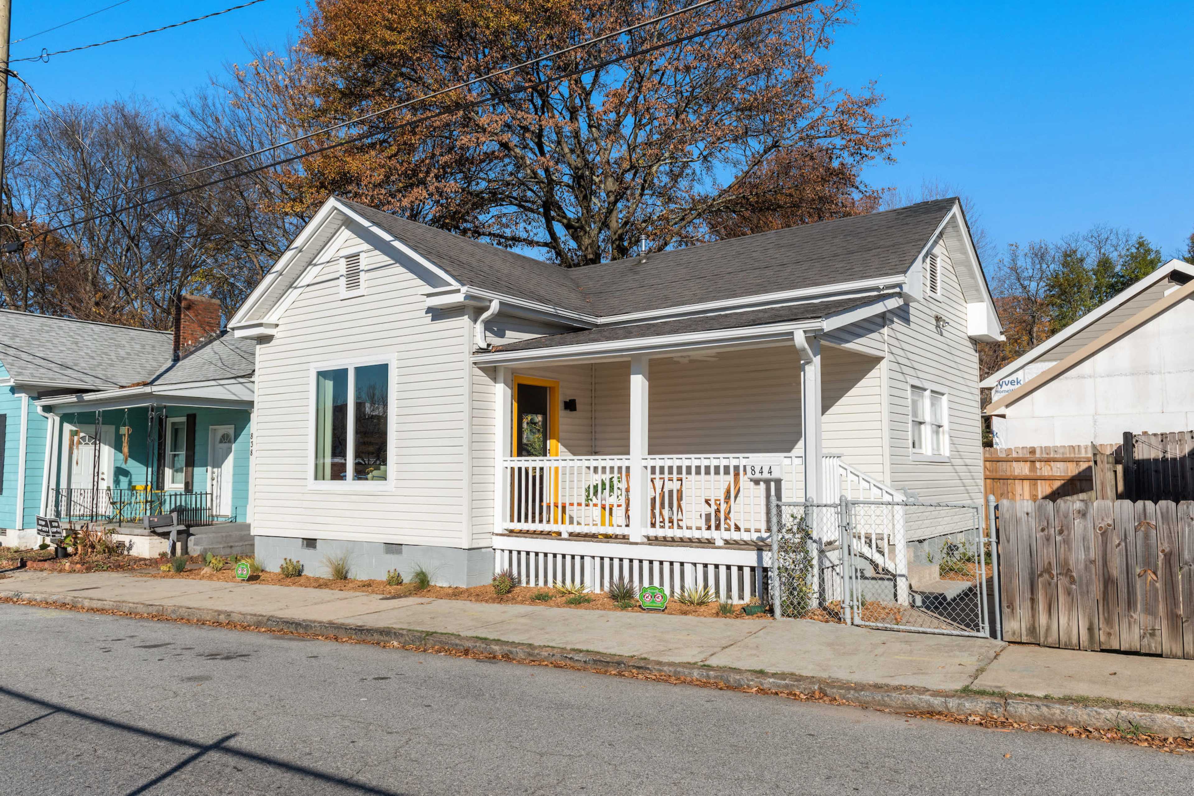 A single-story house with a porch and yellow door is situated on a quiet street, surrounded by trees and neighboring homes.