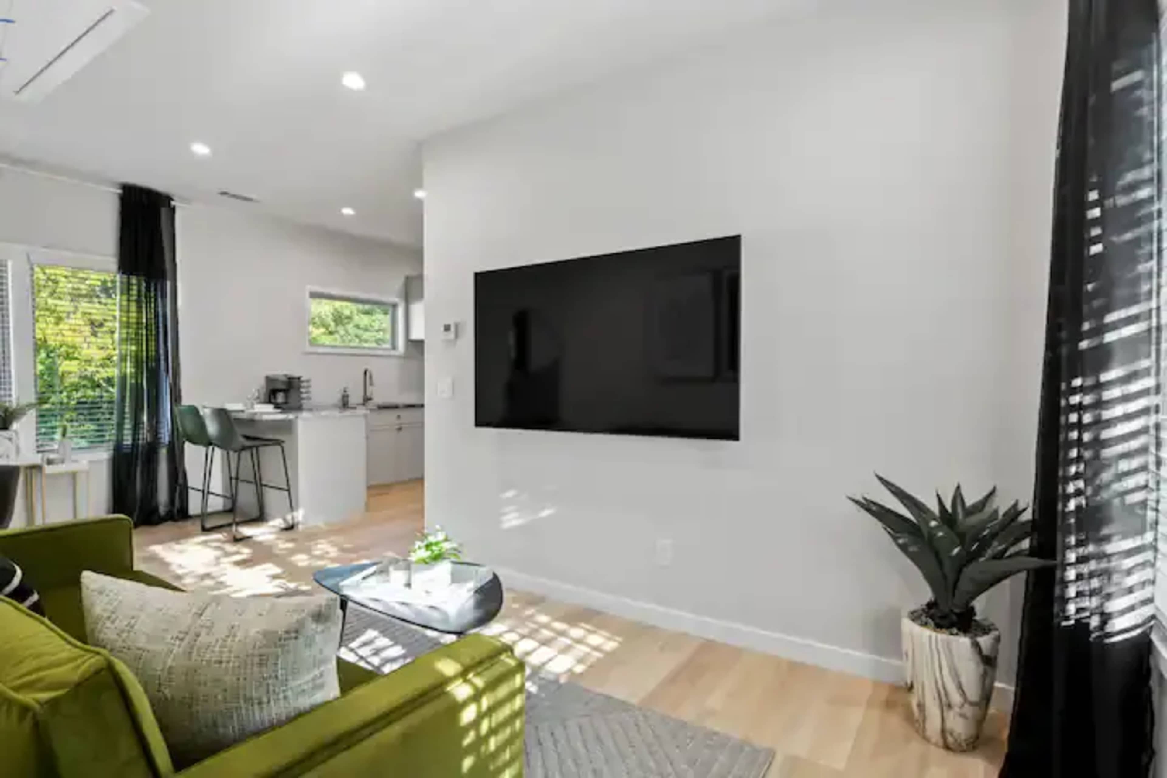 The scene features a modern living space with a mounted television on a light-colored wall, a green sofa, a small coffee table, and large windows allowing natural light to enter.