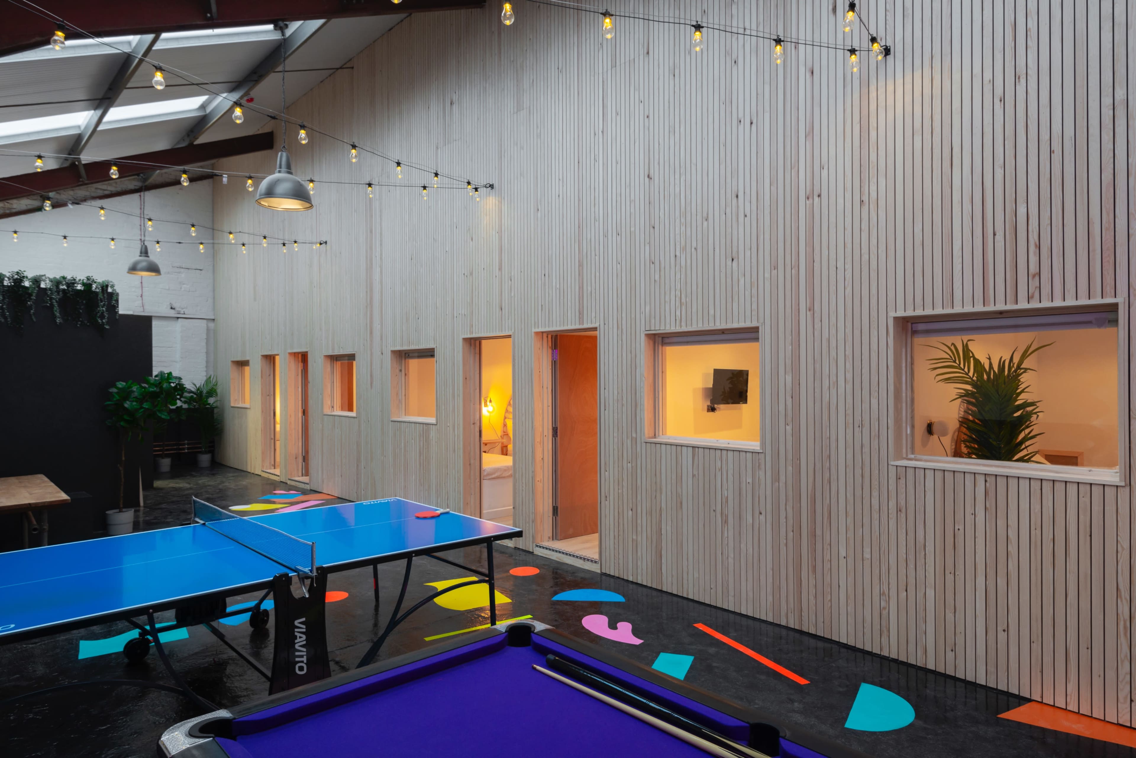 A brightly lit recreational space featuring blue ping pong tables and colorful floor elements, complemented by wooden walls and large windows.