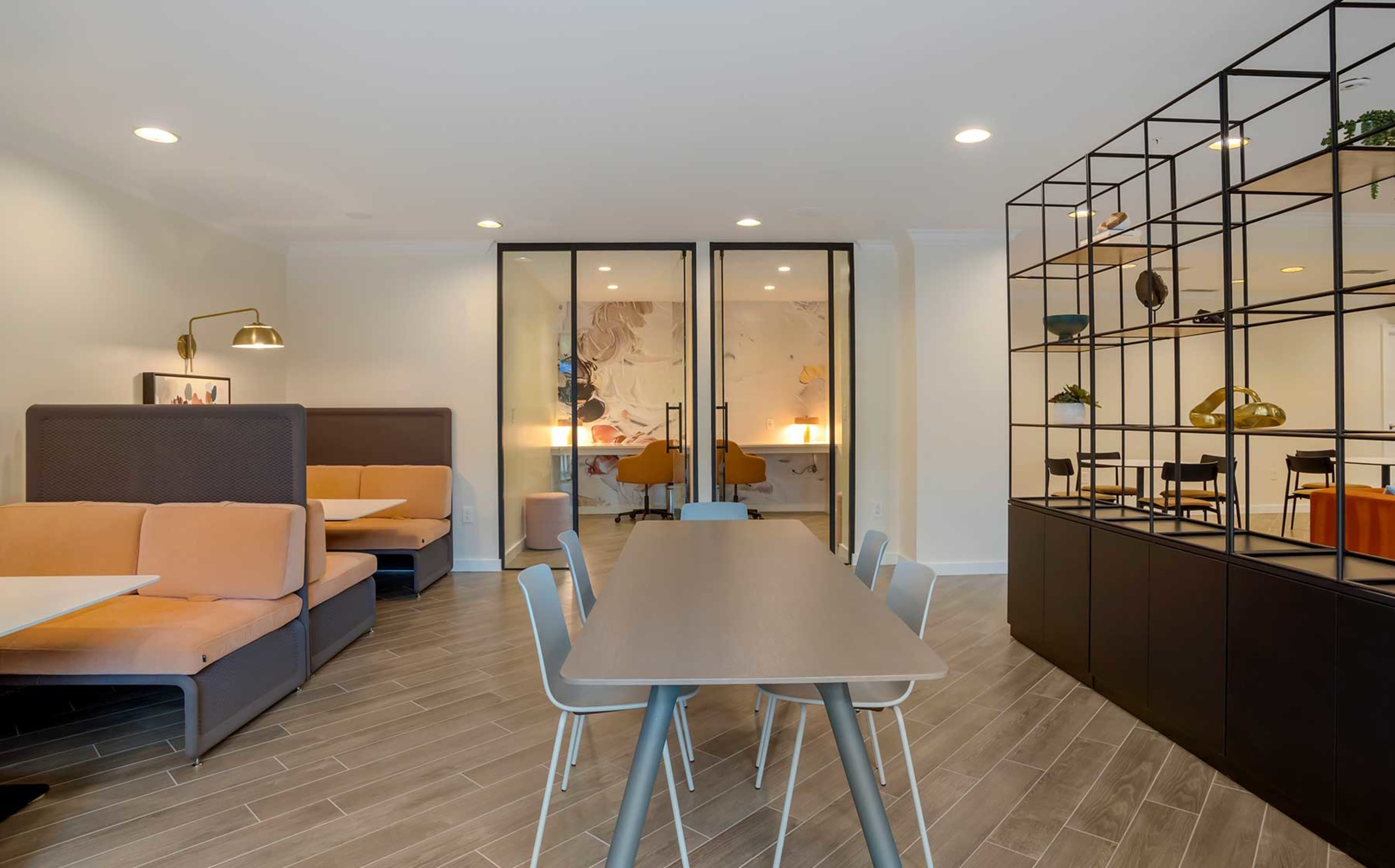 A modern communal space featuring a long table with chairs, soft seating areas, and partitioned workspaces behind glass doors.