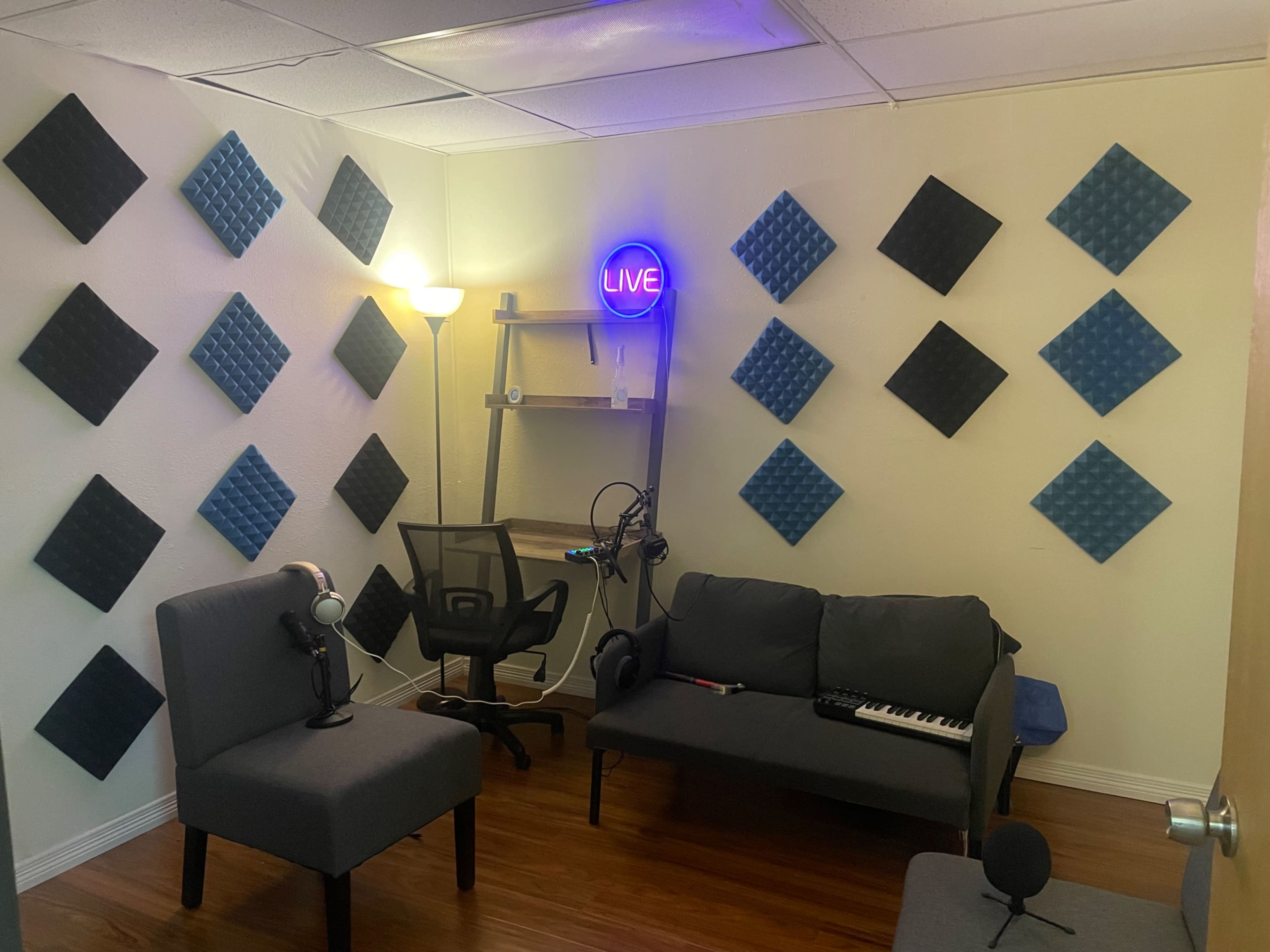 The image shows a small studio room with soundproofing panels on the walls, a black chair, a gray sofa, a microphone setup, and a neon "LIVE" sign.