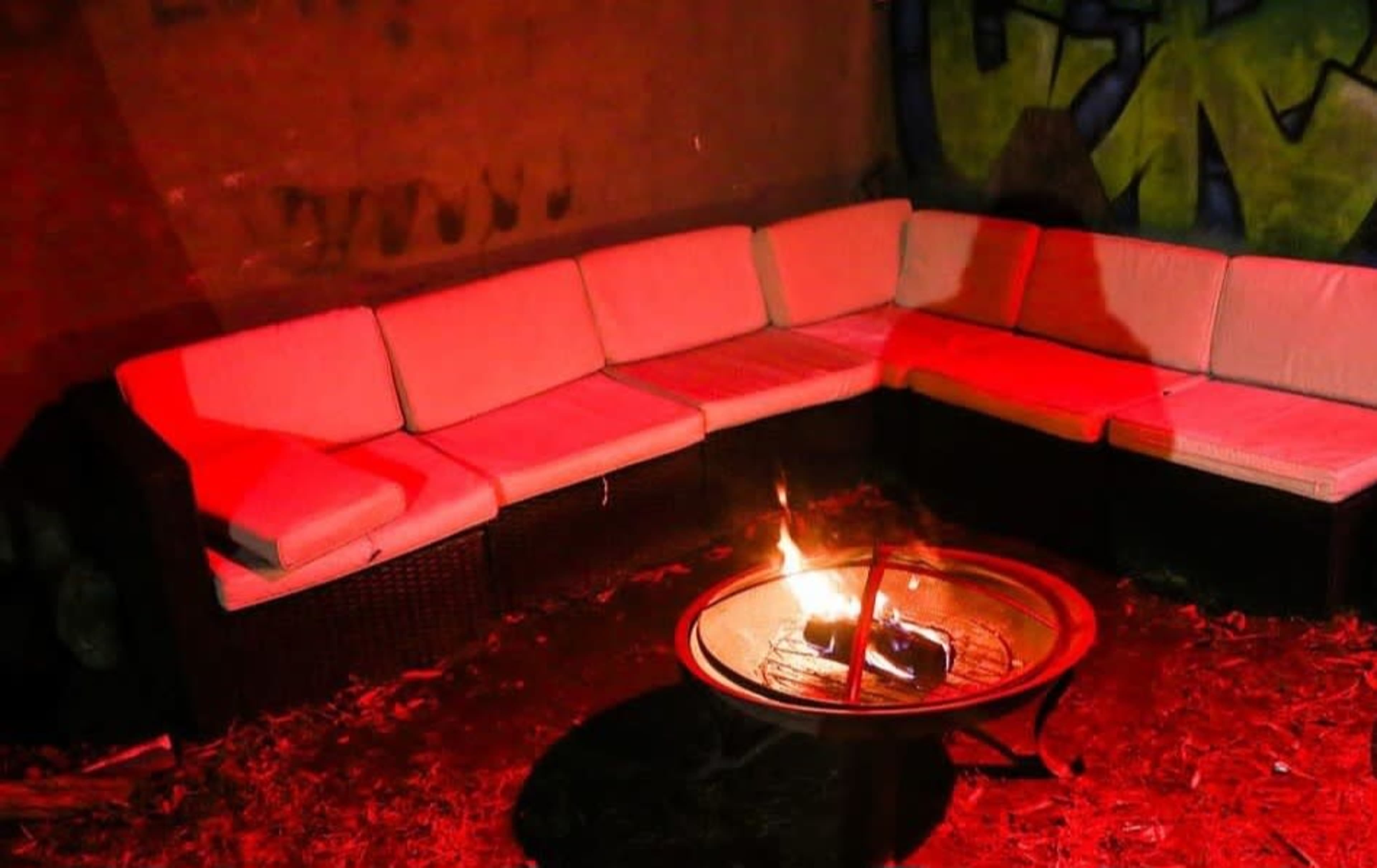 A circular seating area made of white cushions surrounds a central fire pit with flames, all illuminated by red light against a graffiti-covered wall.