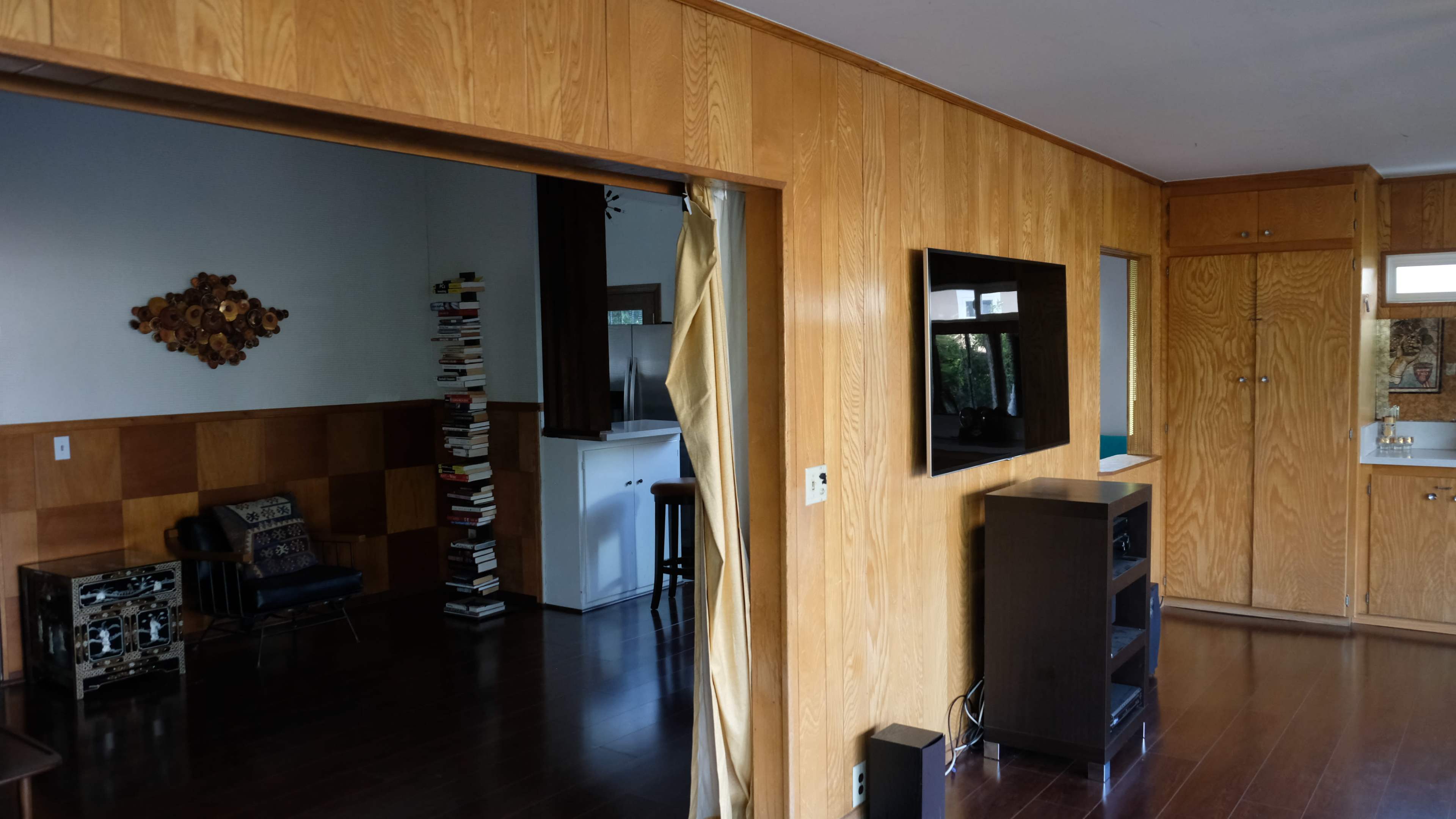 The image shows a room with wooden paneling, a television mounted on the wall, and a doorway leading to another area with a stack of books and a chair.