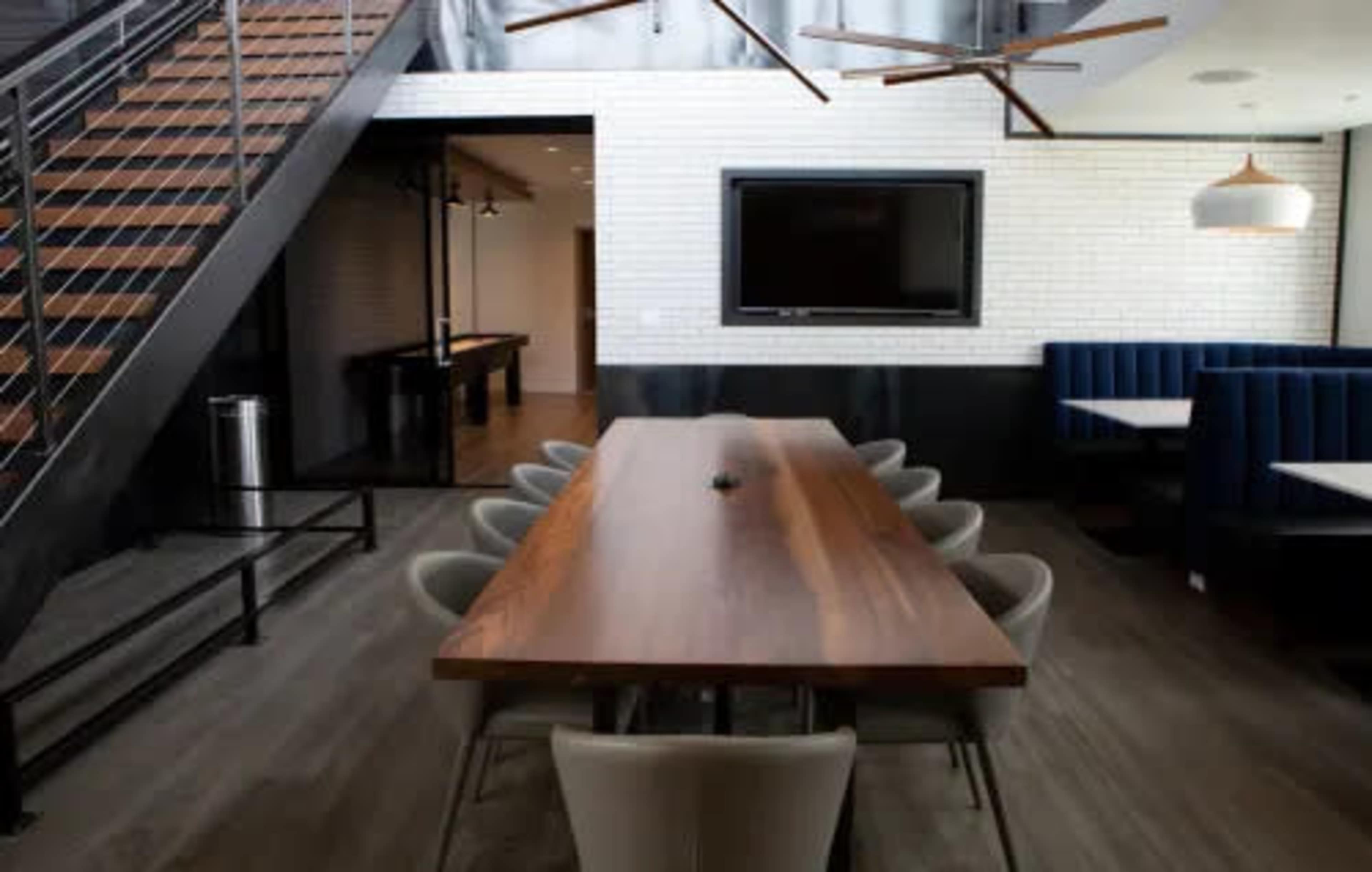A modern dining area with a large wooden table surrounded by gray chairs, set against a backdrop of an open staircase and a seating nook.