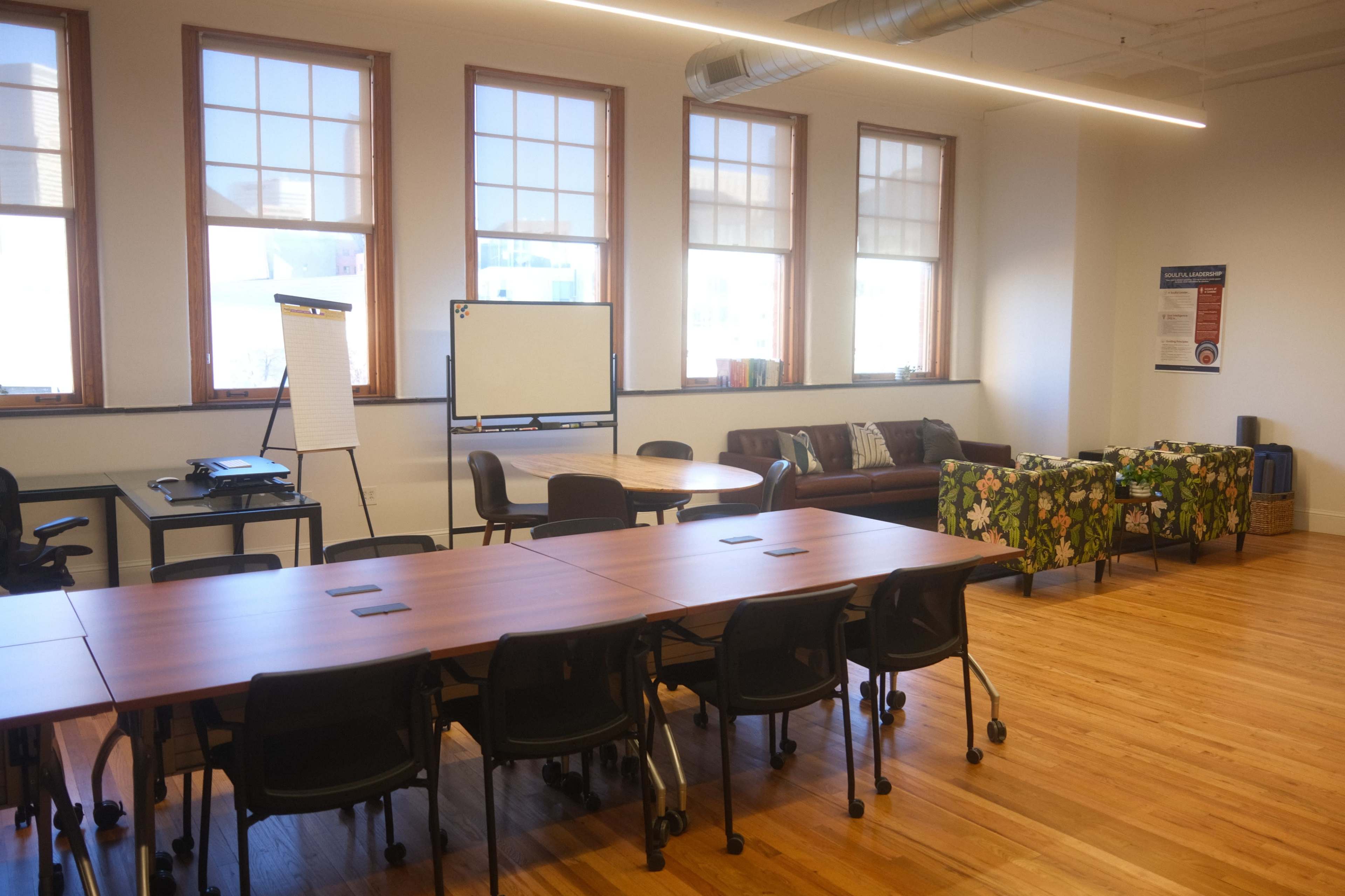 The image shows a modern office space with large windows, a circular table, a sofa covered in floral fabric, and several chairs arranged around adjustable desks.