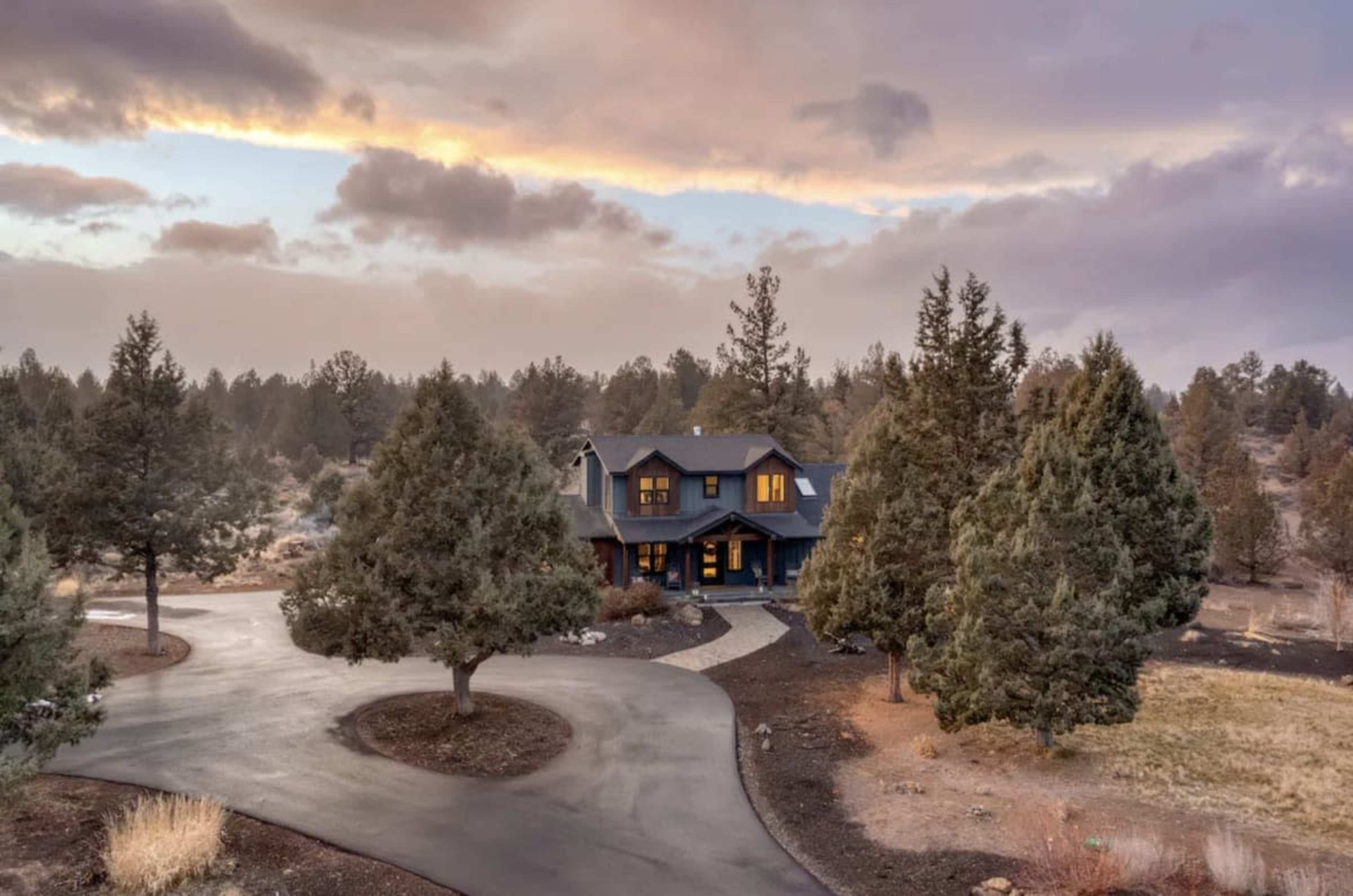 A dark blue house with a winding driveway is surrounded by evergreen trees against a cloudy sky at dusk.