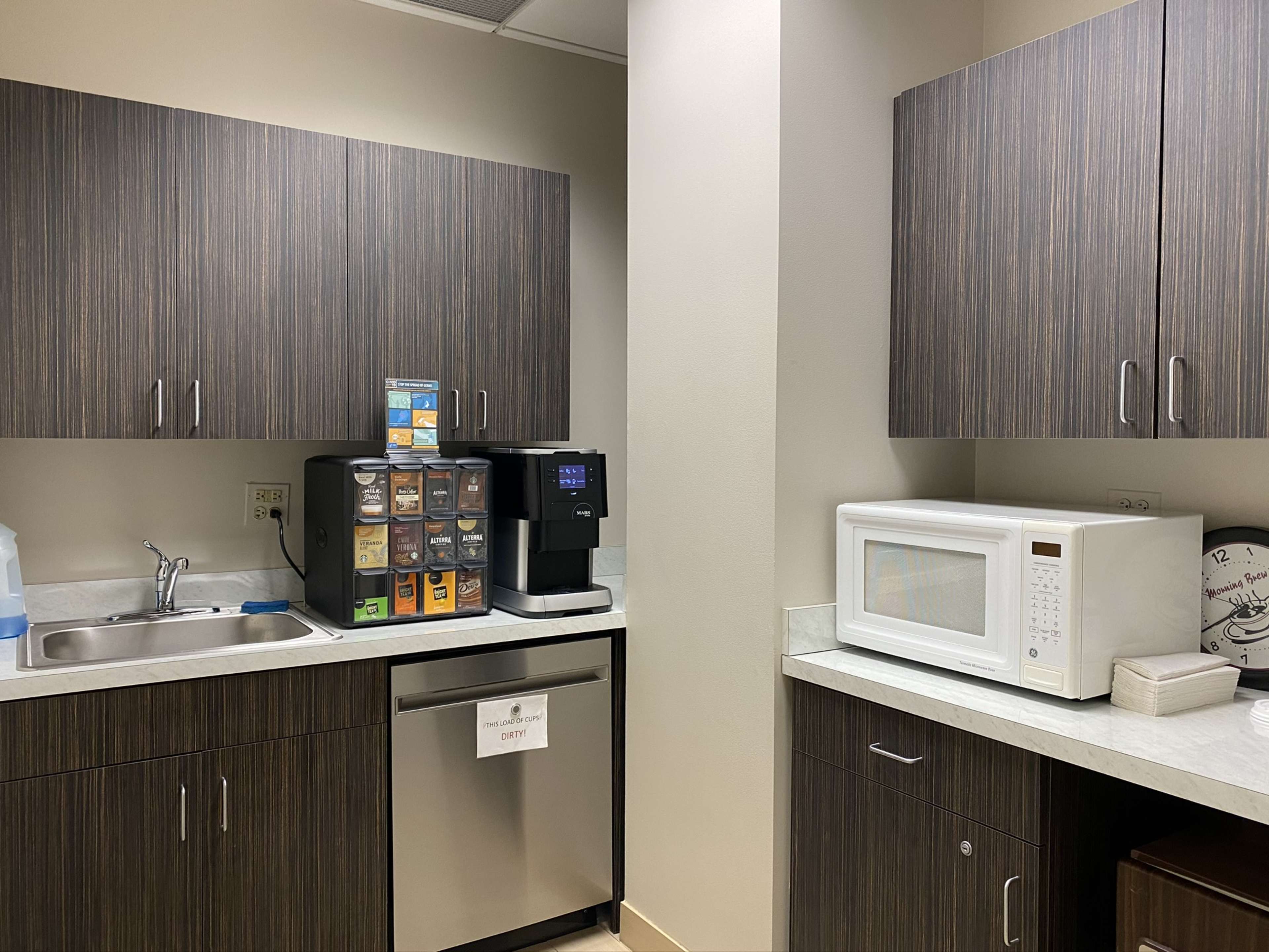 The image shows a break room with dark wooden cabinets, a coffee machine, a beverage dispenser, a microwave, and a sink.