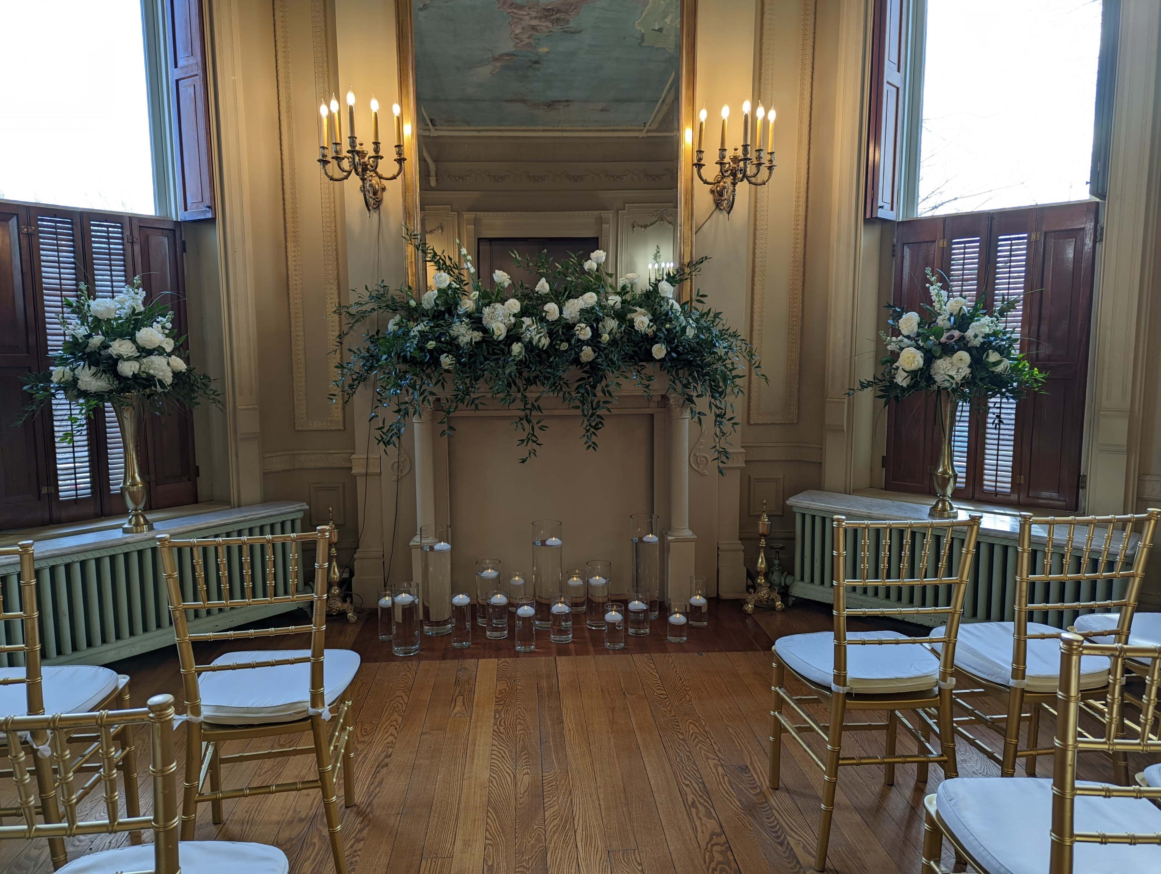 The image shows a decorated indoor wedding setting with chairs arranged in front of a floral-adorned mantelpiece, illuminated by candlelight.