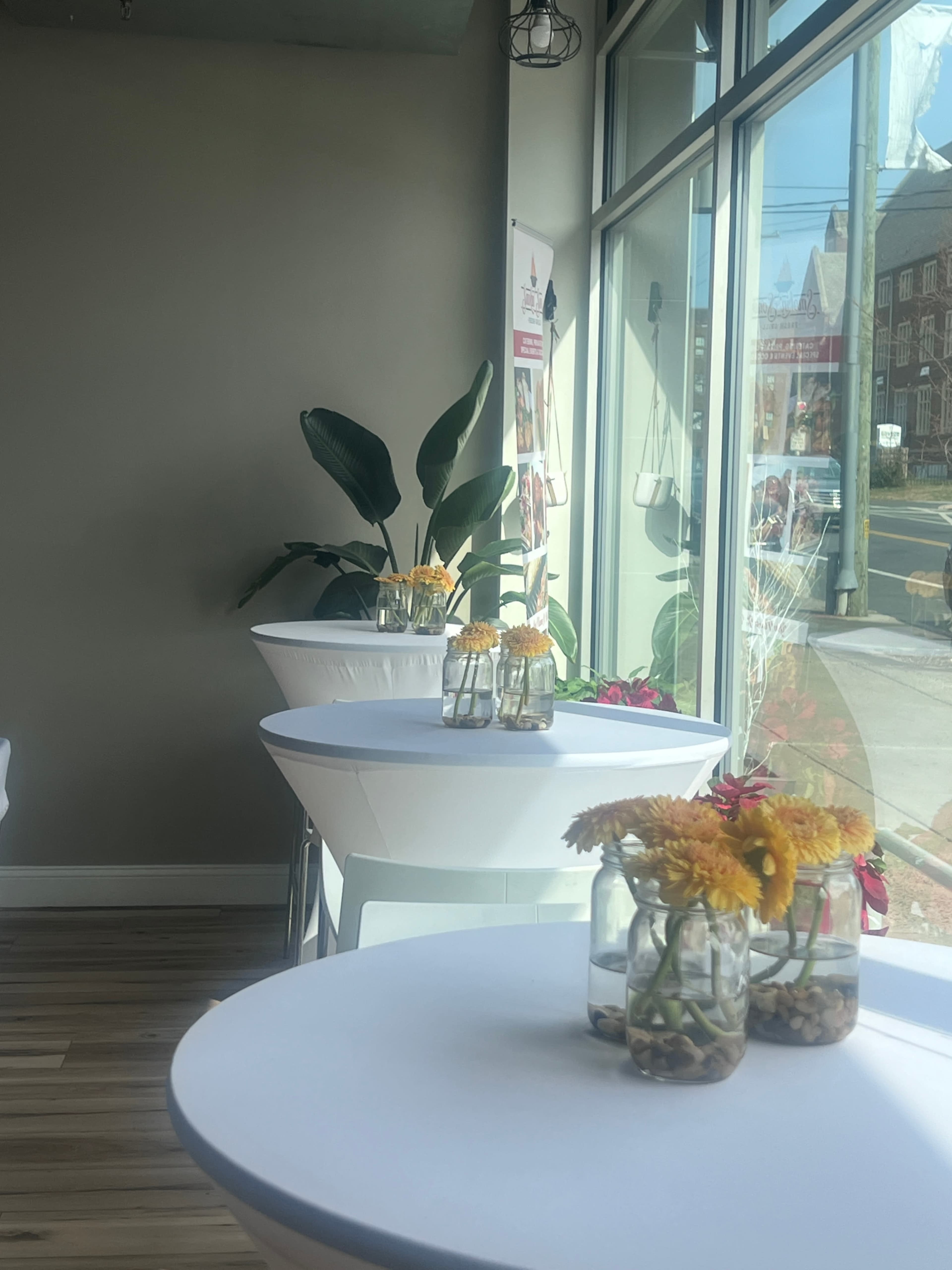 The image shows a bright interior with several round tables adorned with flowers in glass vases and potted plants near the window.