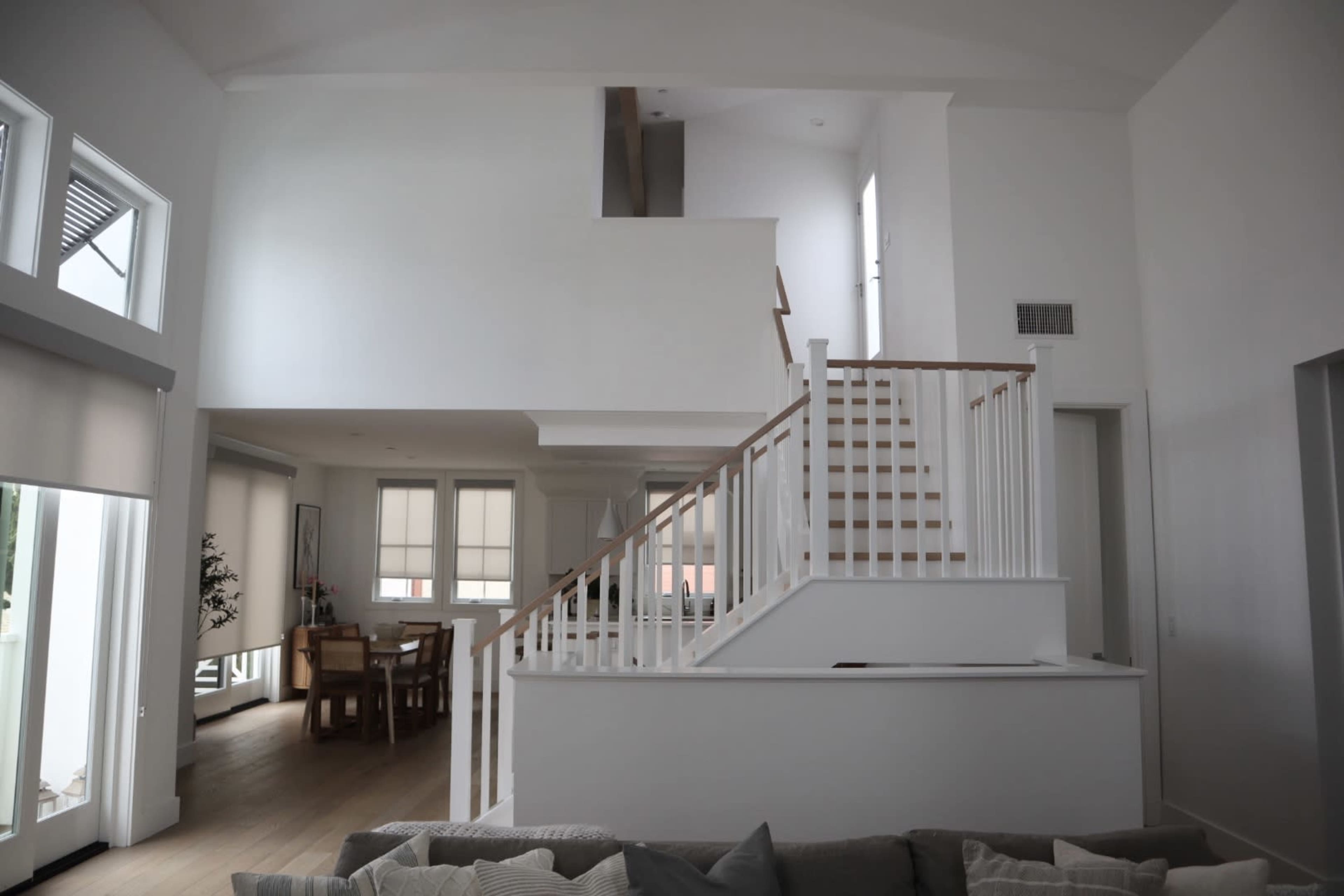 A modern interior of a house with a staircase leading to an upper level, a dining area in the background, and large windows allowing natural light.