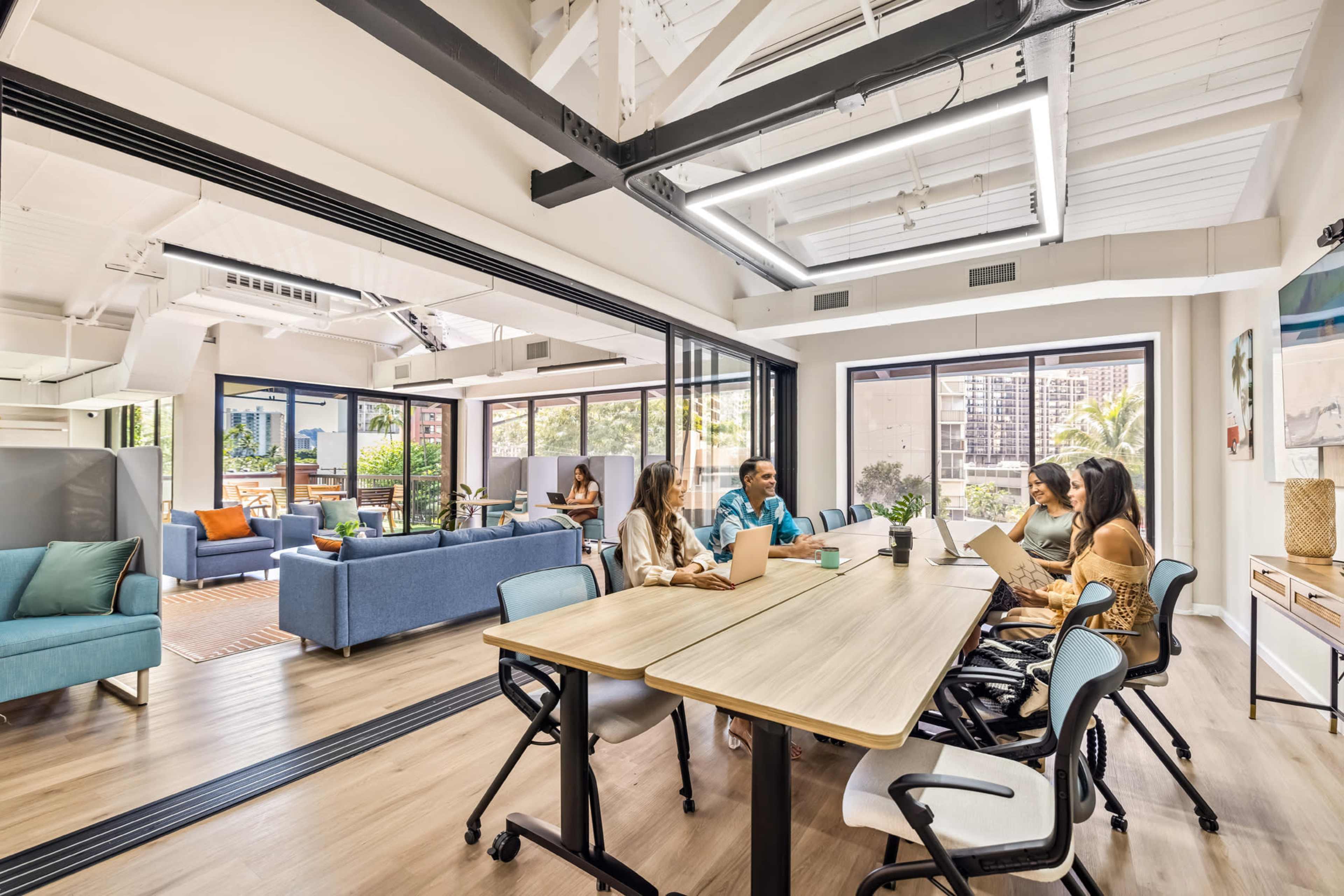 A group of four people sit around a large table in a modern office space with large windows and a lounge area.