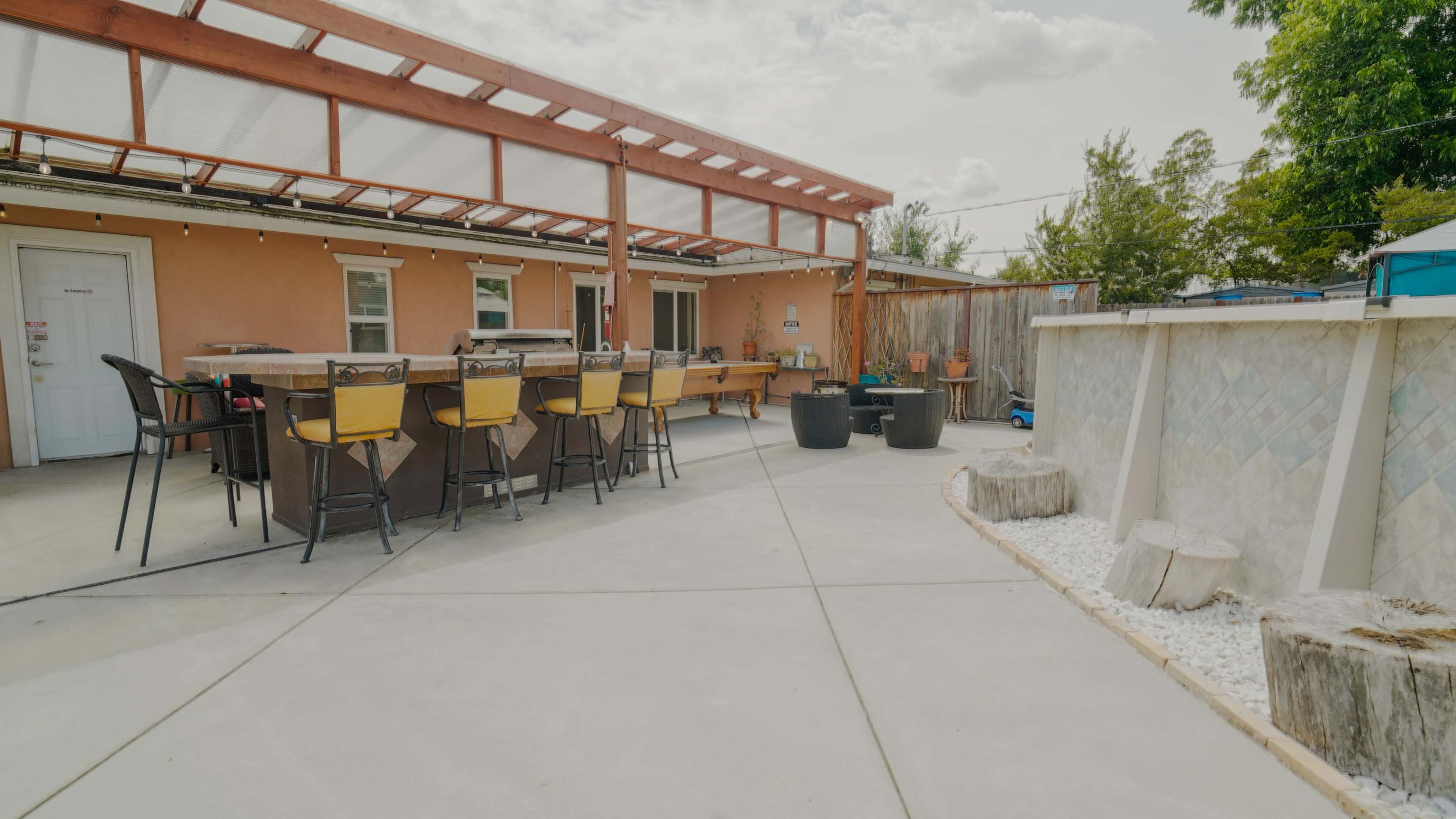 The image shows an outdoor patio area featuring a bar with high chairs, a fence, and decorative landscaping elements.