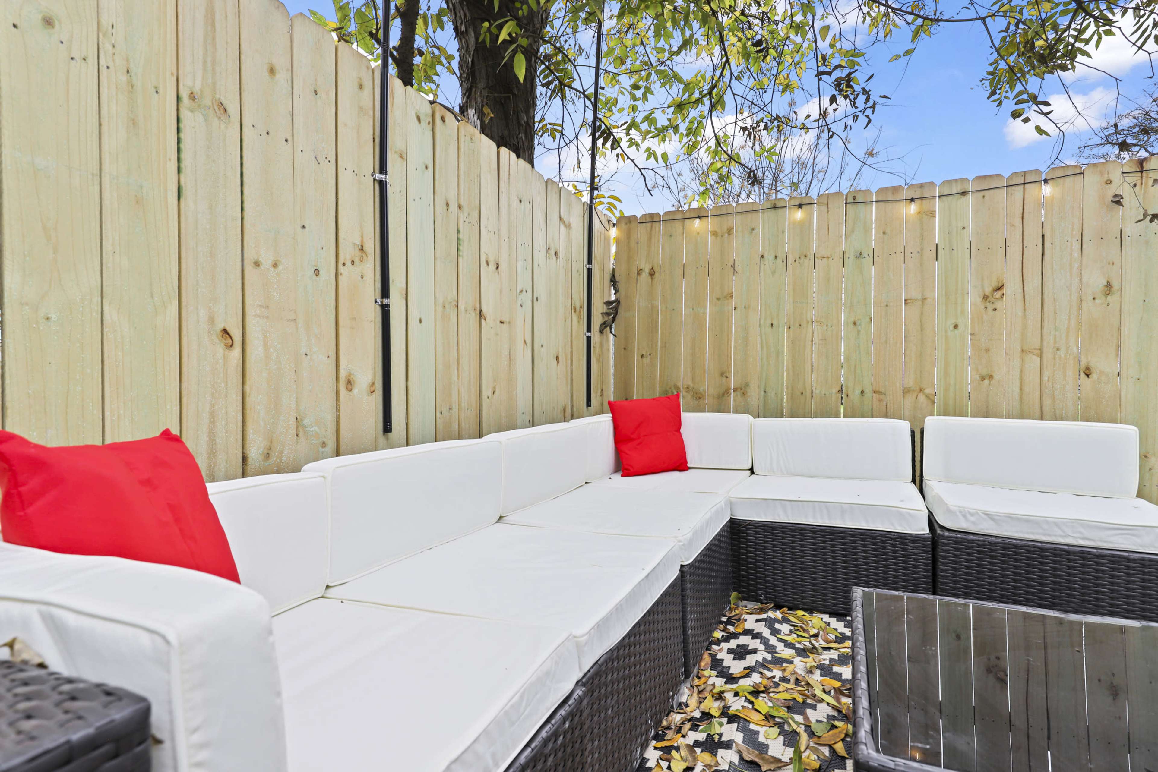 The image shows a patio area with a white outdoor sectional sofa, red throw pillows, and a glass-top coffee table, enclosed by a wooden fence and surrounded by fallen leaves.