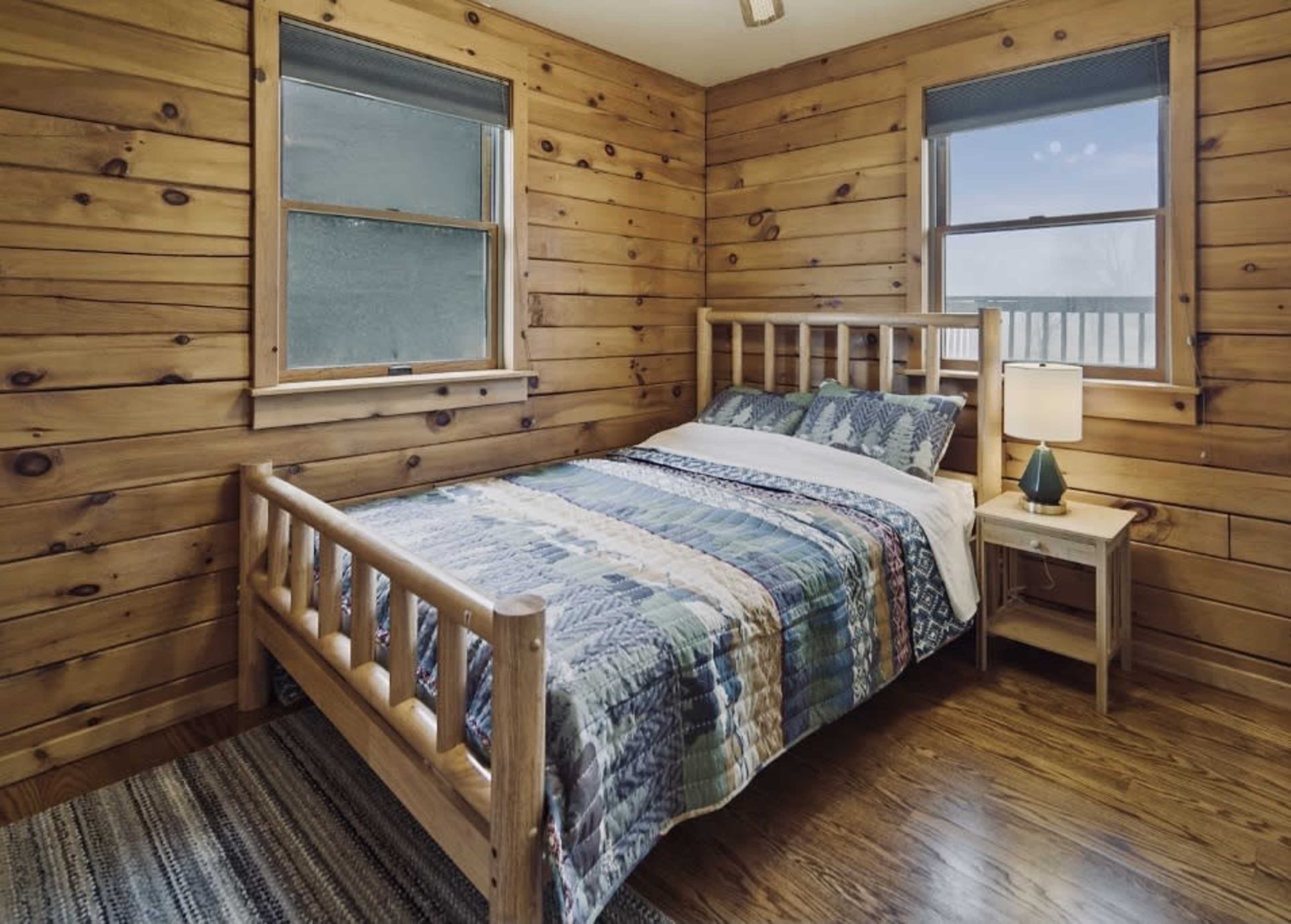 A wooden-paneled bedroom features a bed with a patchwork quilt, two windows, and a nightstand with a lamp.