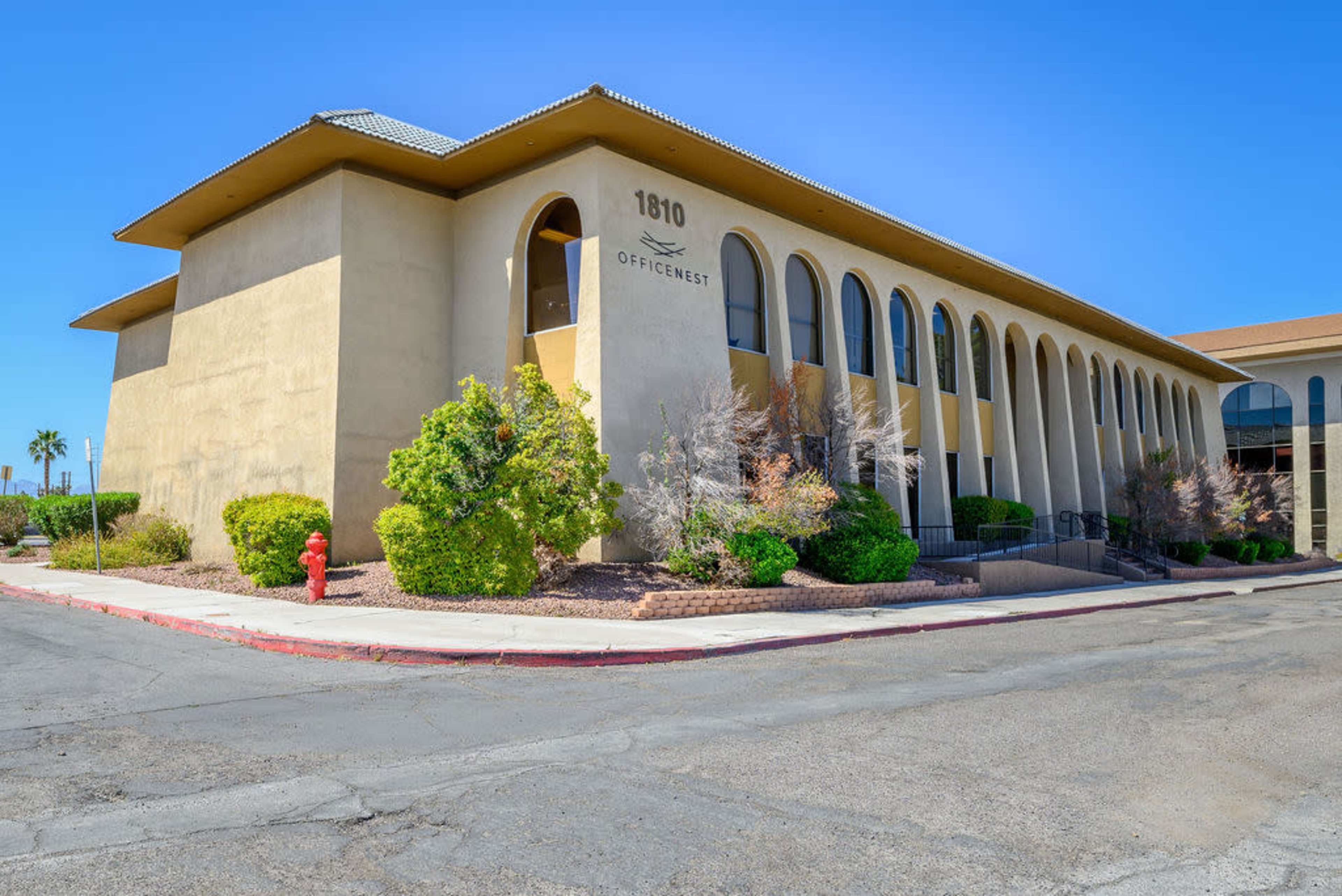 The image shows a two-story office building with arched windows, situated on a street corner and surrounded by landscaped shrubbery.