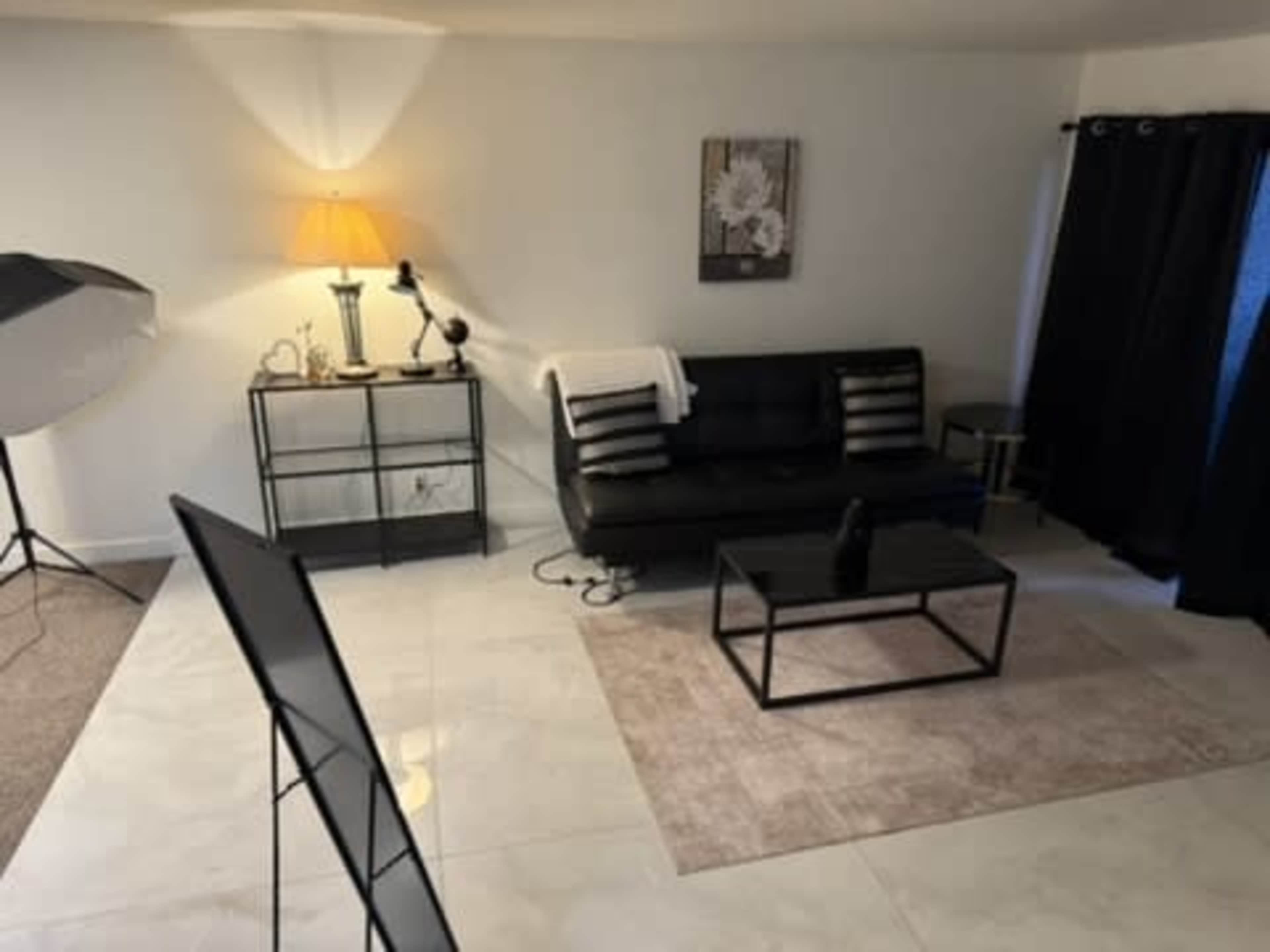 The image shows a modern living room with a black sofa, a coffee table on a light rug, a lamp, and a dark curtain covering a window.