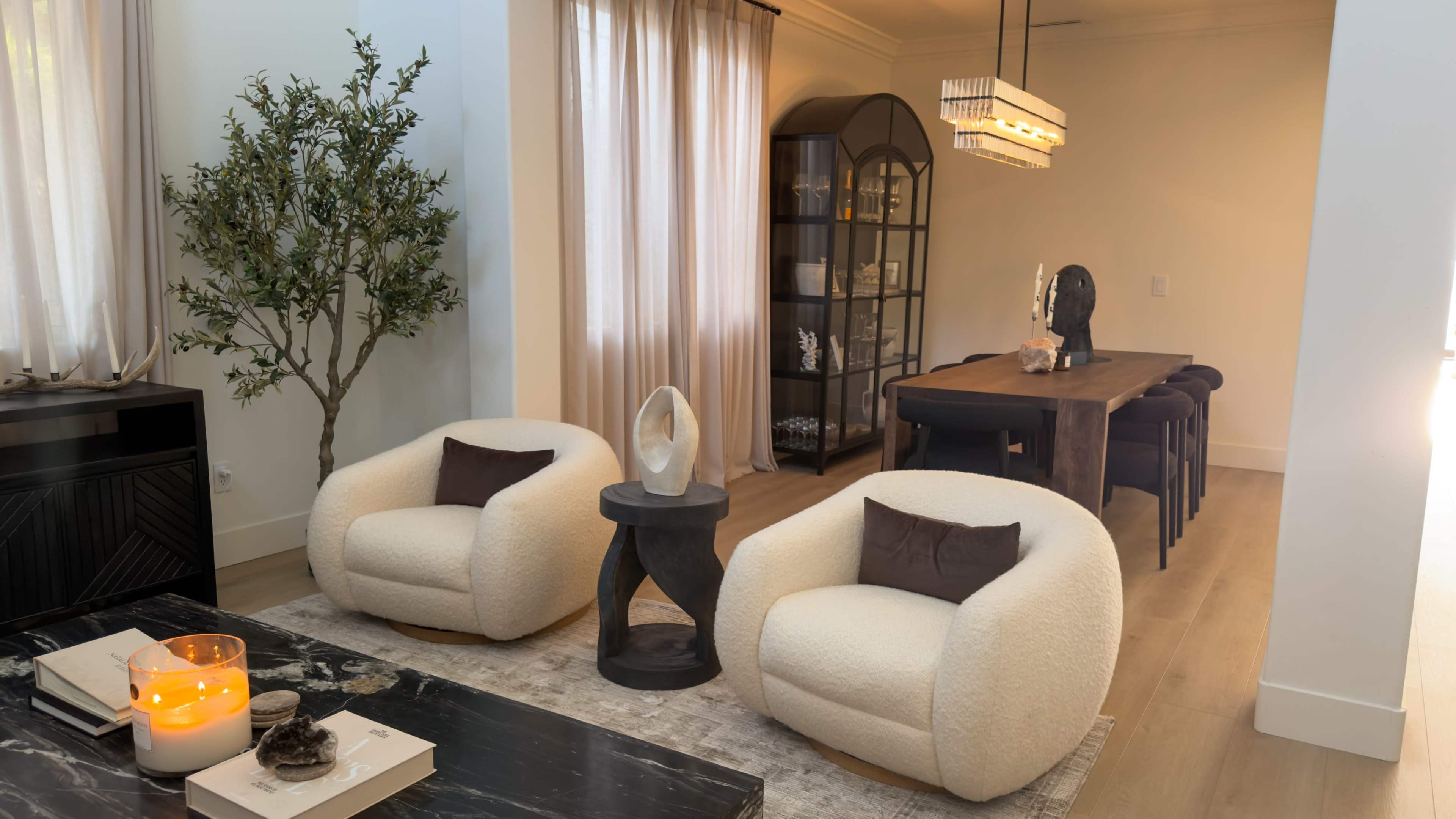 The image shows a modern living room featuring two plush white chairs, a small black side table, an olive tree, and a dining area with a long wooden table and elegant light fixture.
