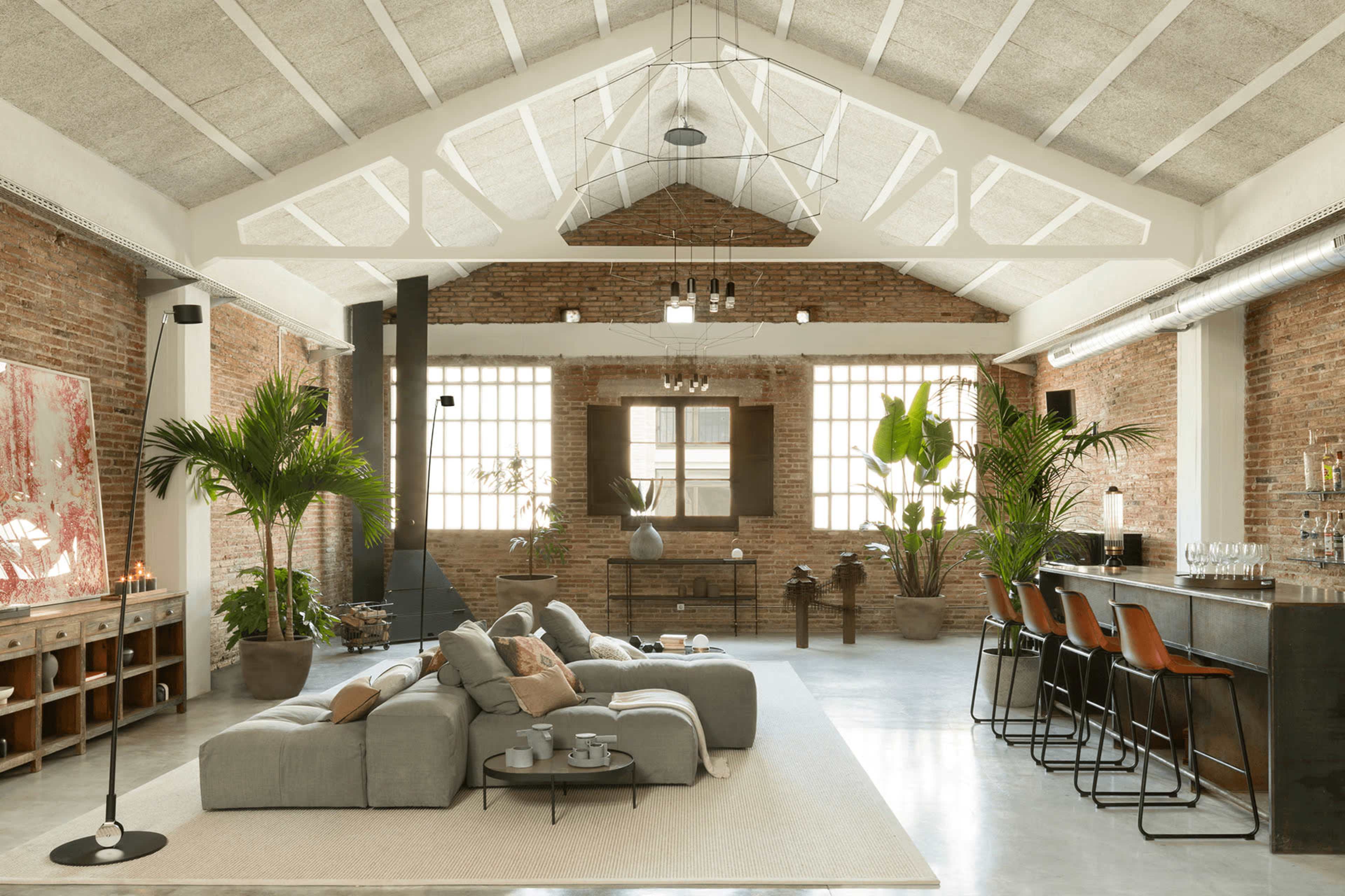 The image shows a spacious loft interior with high ceilings, exposed brick walls, a comfortable seating area, and a bar with stools, adorned with tropical plants.