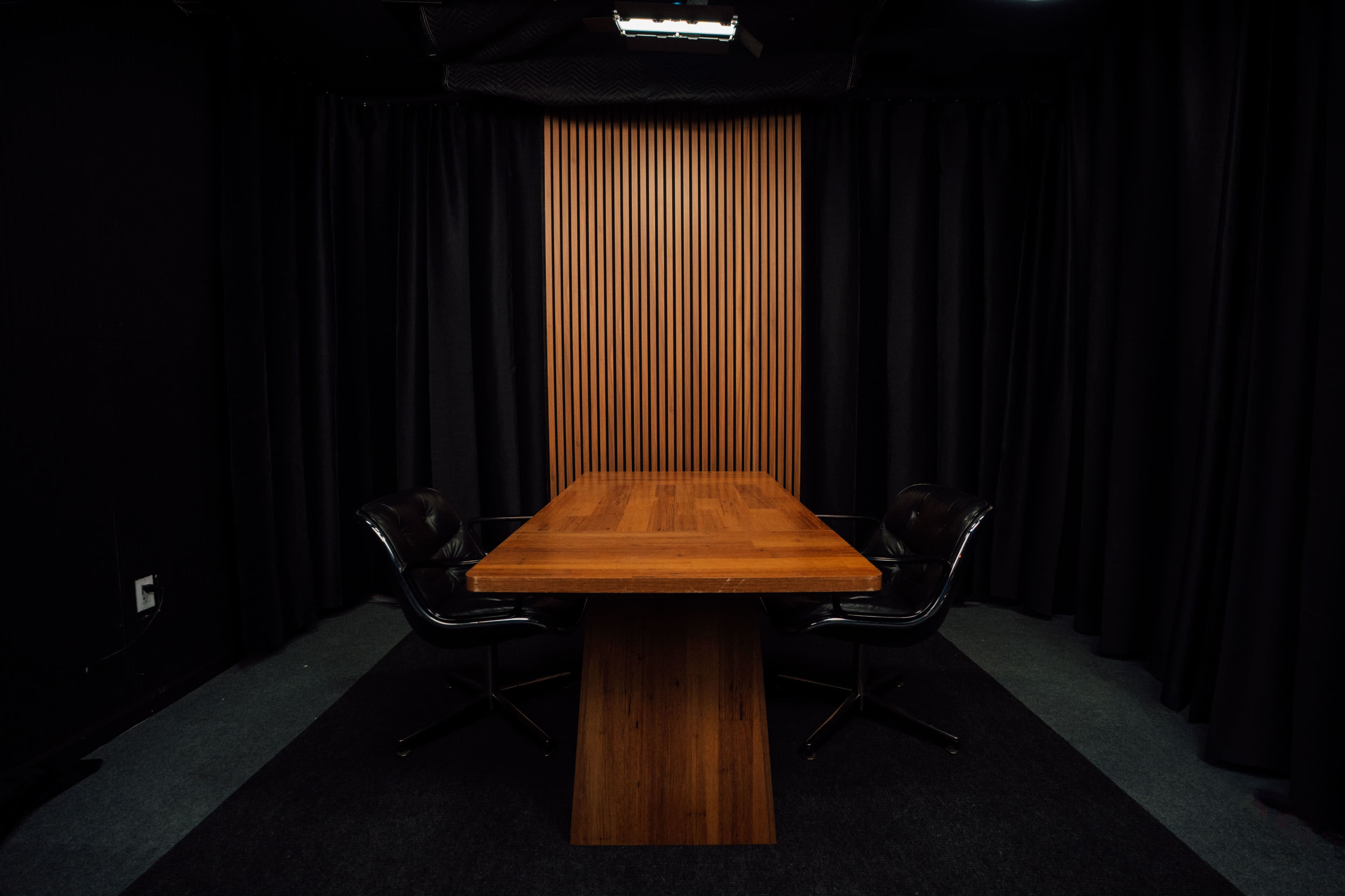 The image shows a minimalist conference room with a wooden table and two black leather chairs, surrounded by black curtains.