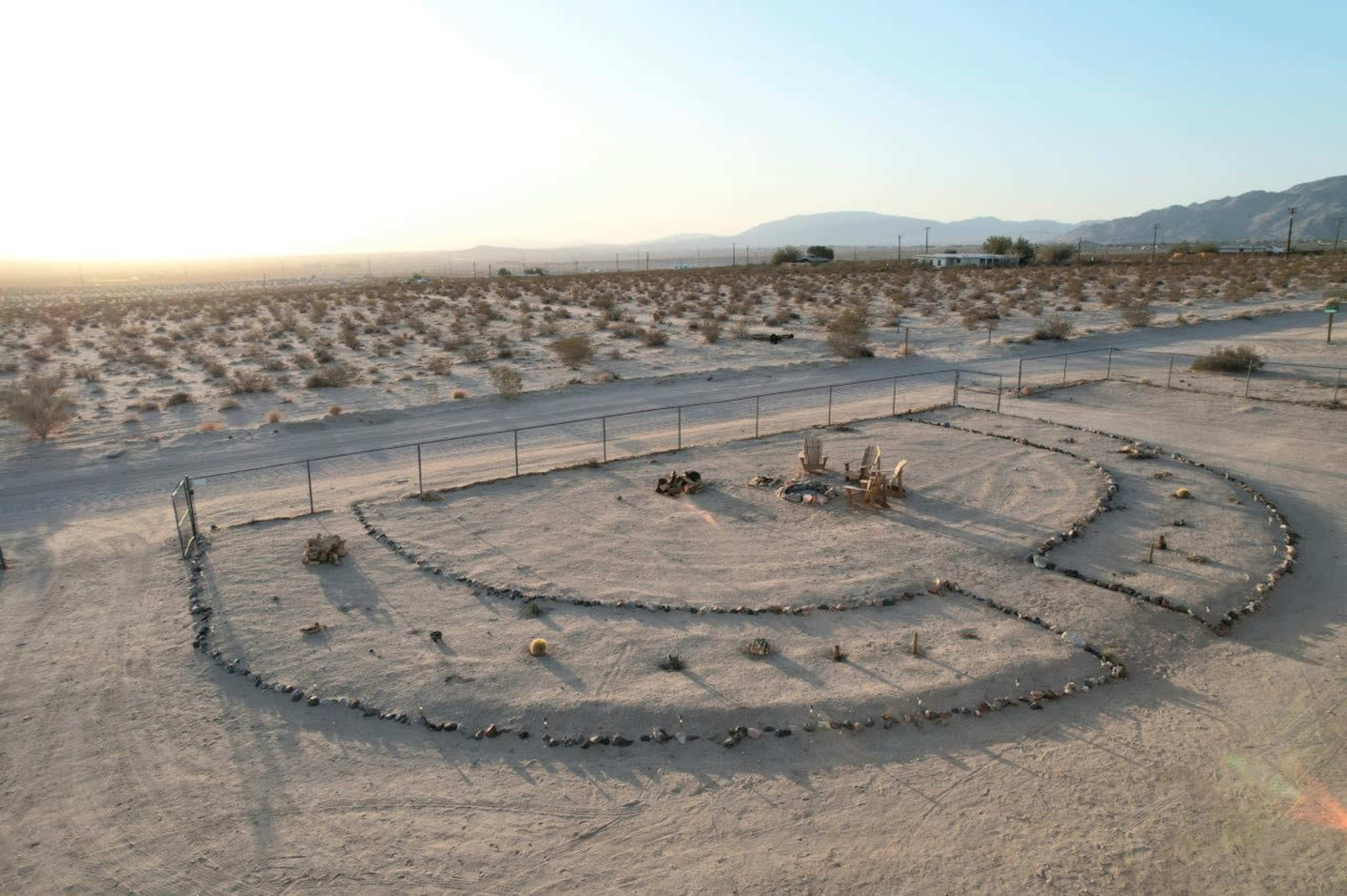 The image shows a circular stone arrangement in a desert landscape, surrounded by sparse vegetation and mountains in the background.