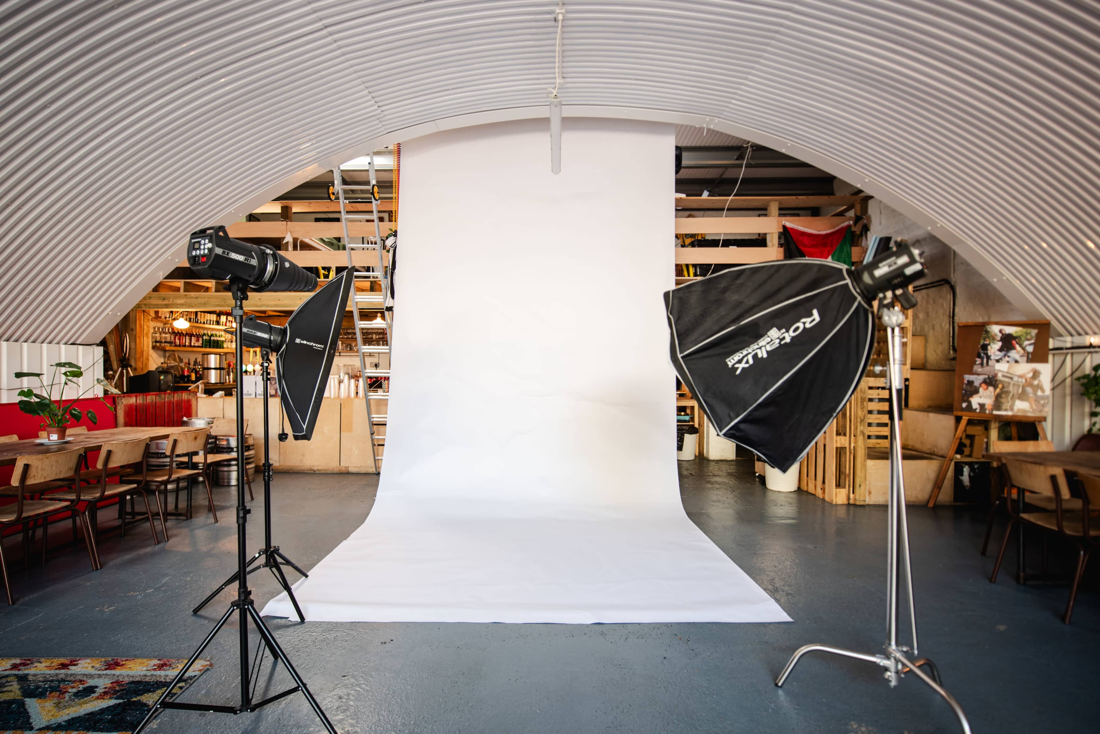 The image shows a photography studio with a white backdrop, studio lights on tripods, and a spacious interior featuring wooden shelves and furniture.