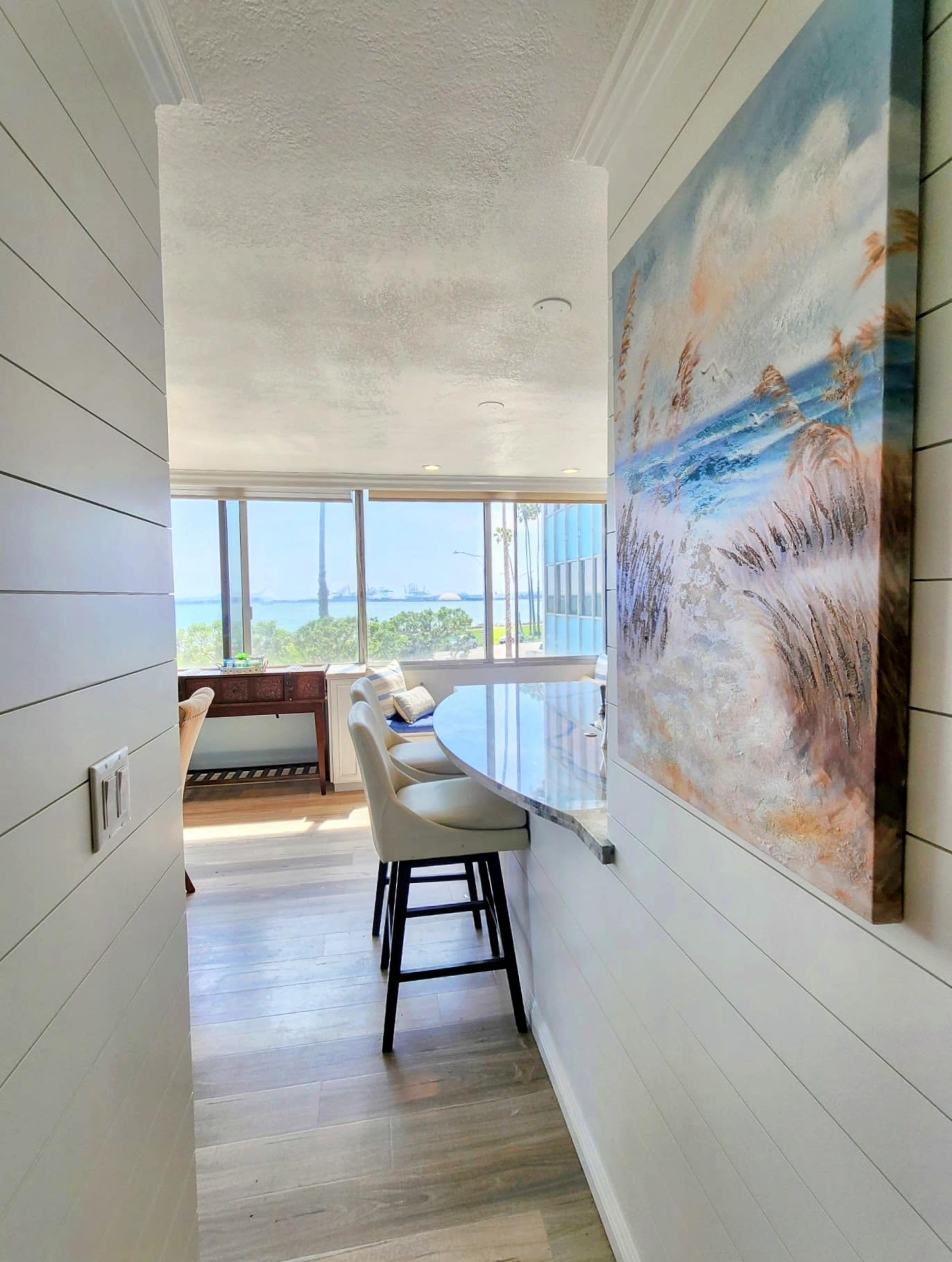 A hallway with white paneling leads to a bright room featuring large windows and a view of the ocean, decorated with a painting of a beach scene.