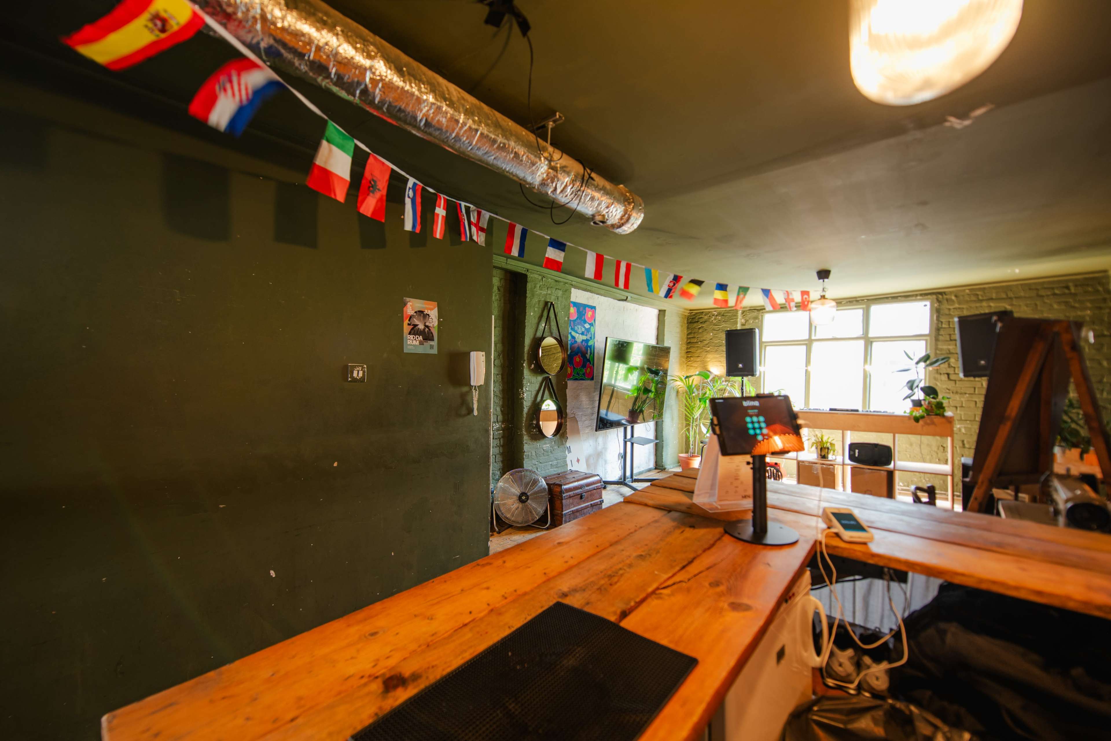The image shows a cozy interior of a cafe or bar with wooden furniture, green walls, and various national flags hanging from the ceiling.